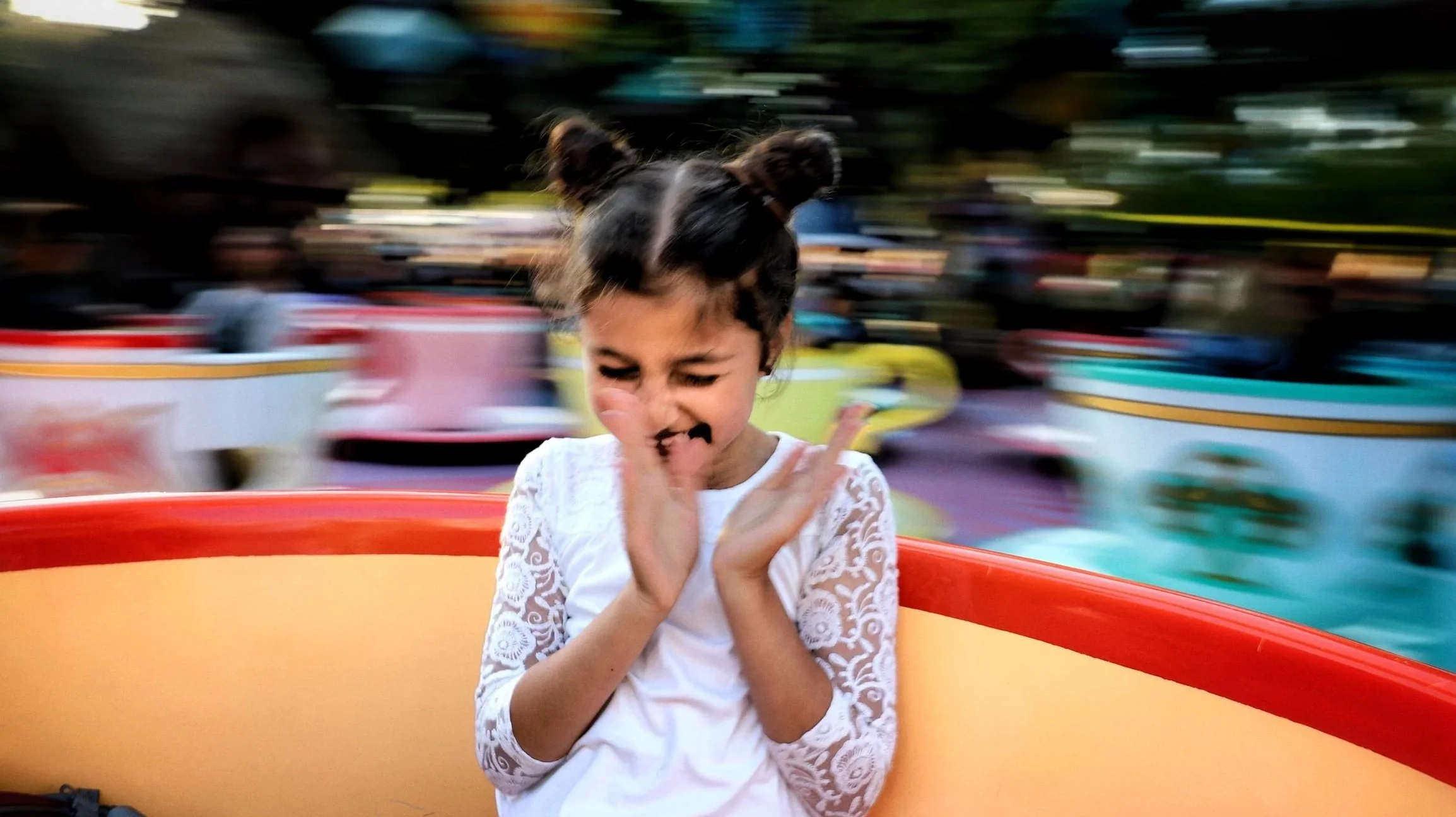 A young girl with dark hair in two buns, wearing a white lace dress, is enjoying a ride on a colorful amusement park ride, with a joyful expression and her hands crossed near her face, while the background shows motion-blurred amusement rides and att