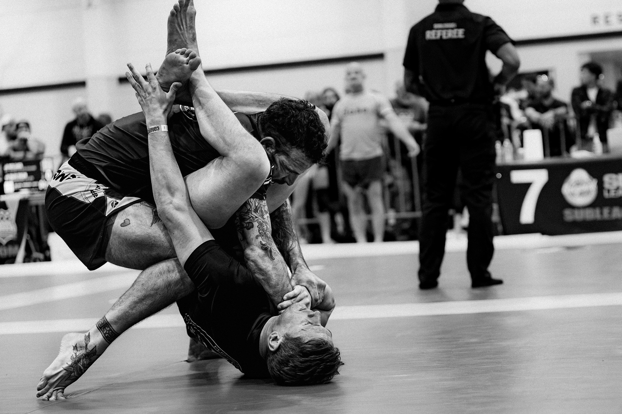 Two martial artists grappling on the mat during a match, with one in a dominant position holding the other in a submission hold, referees and spectators in the background.