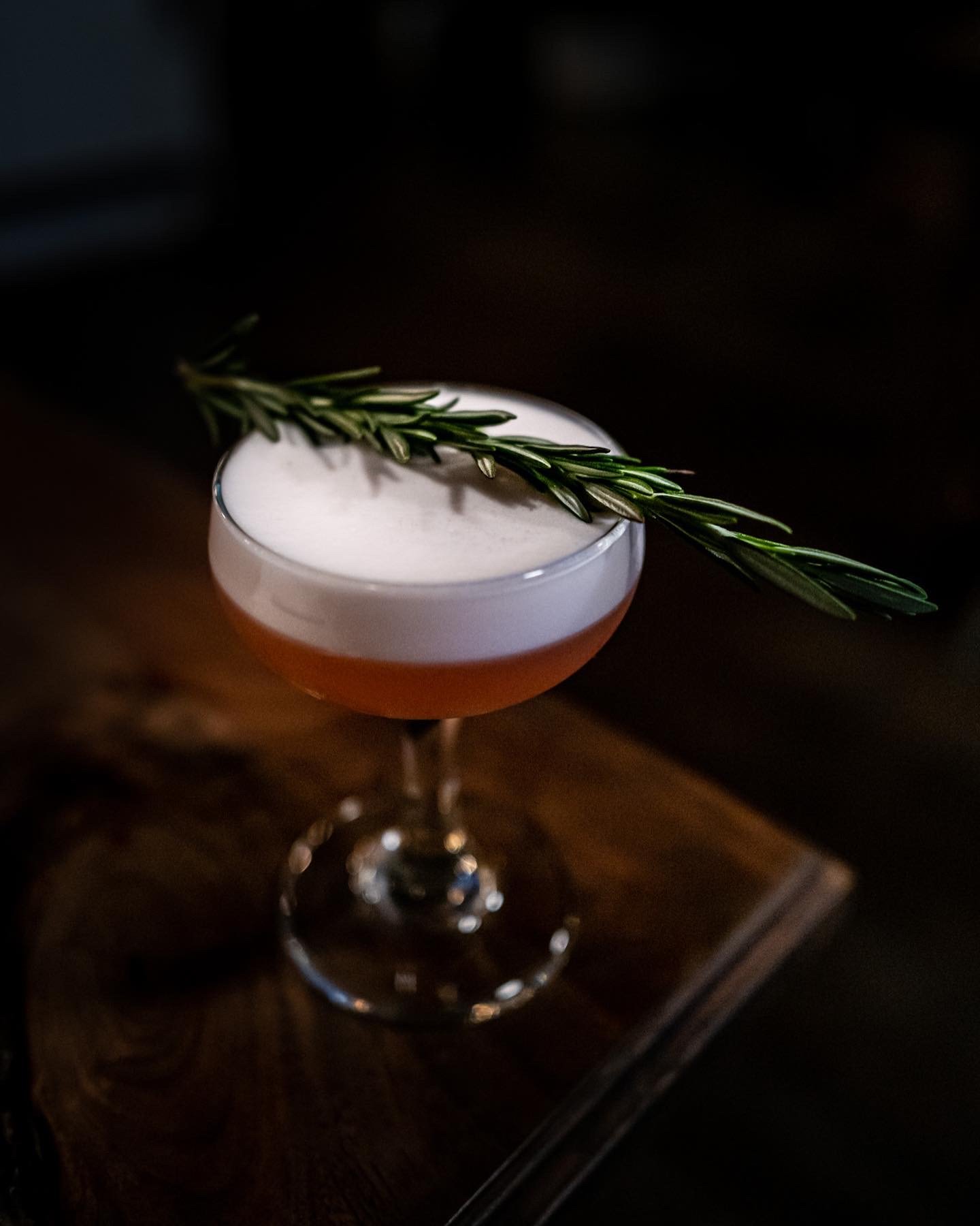 Cocktail glass with foam on top, garnished with a sprig of rosemary on a wooden surface.