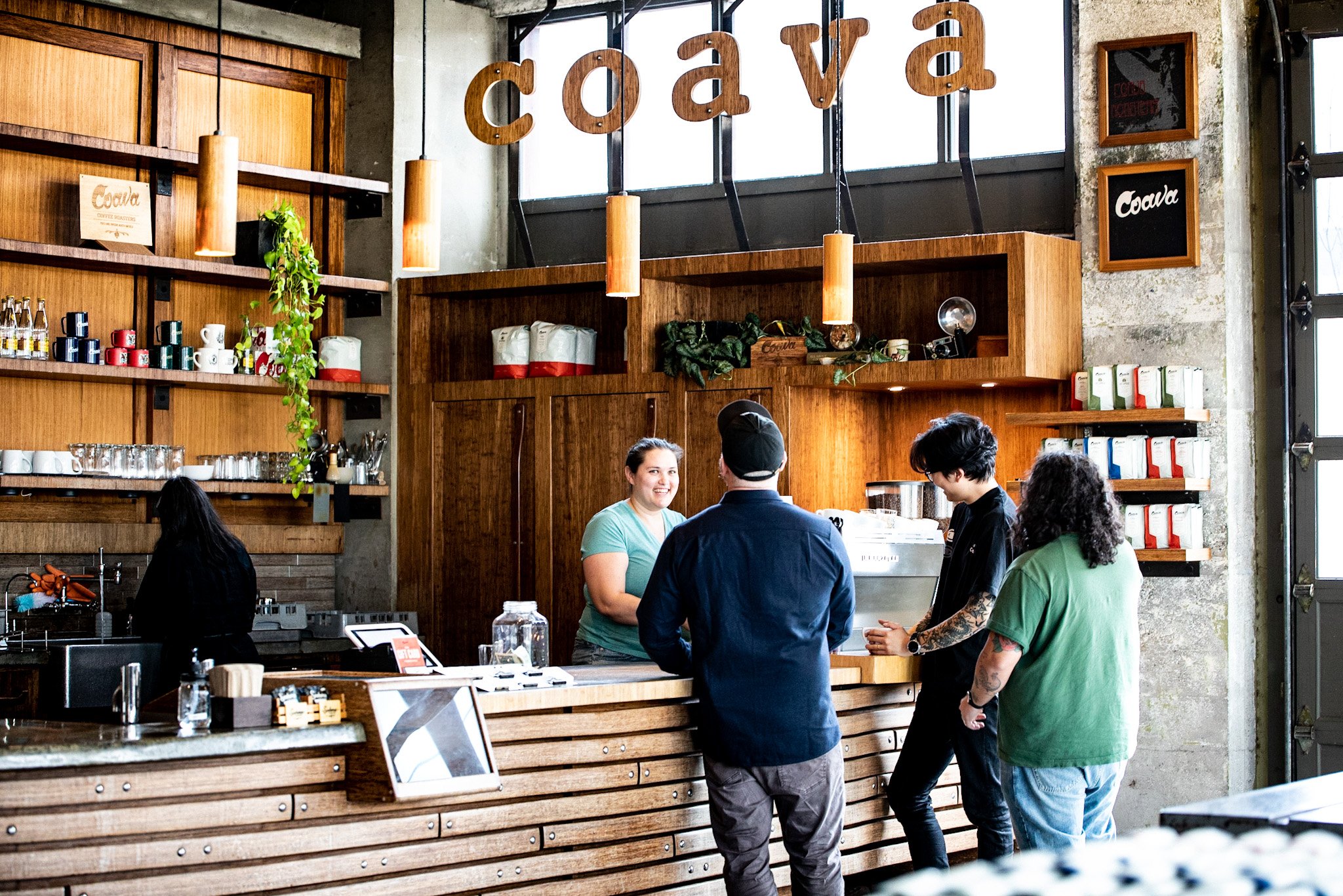 Inside a coffee shop with wooden decor, several customers and staff interacting at the counter, coffee mugs on shelves, and a large sign spelling 'coffee' hanging above.