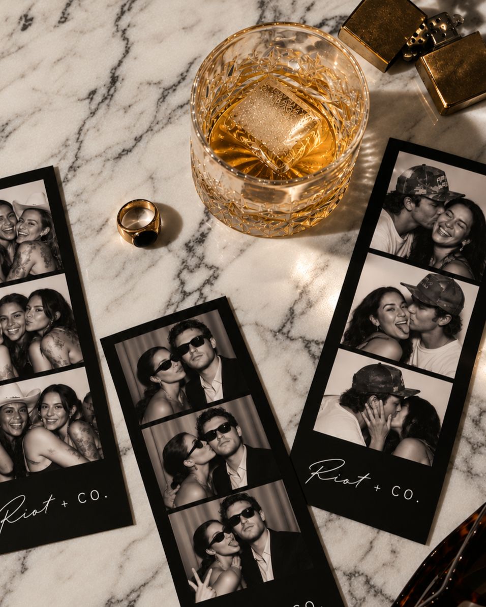 Photo booth contact sheets with black and white images of young couples and friends, featuring playful poses, wearing sunglasses, and romantic moments, placed on a marble surface alongside a glass of whiskey with an ice cube, a gold ring, and two small gold decorative objects.