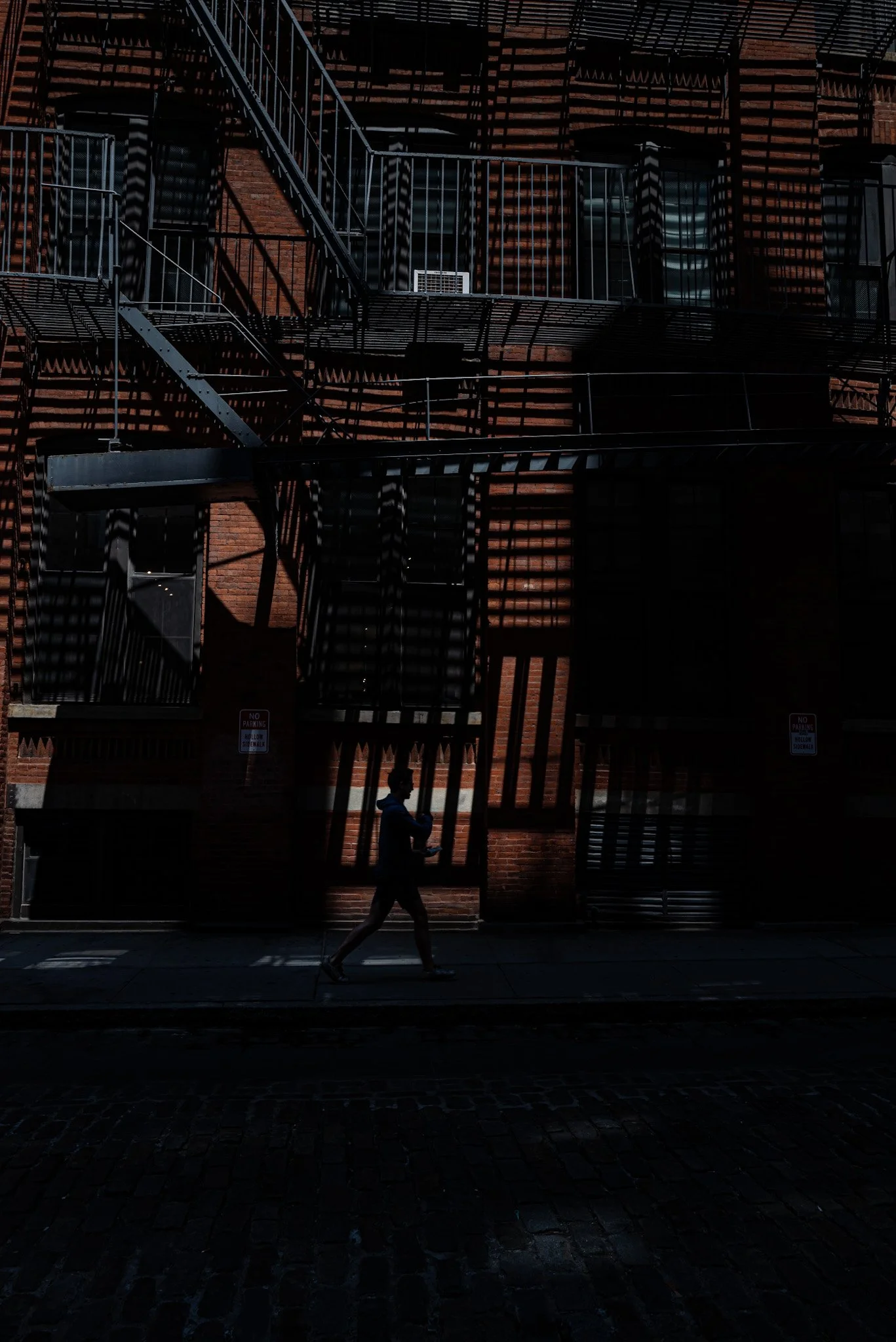 A person walking on a sidewalk in front of a brick building with fire escapes creating shadows on the wall.