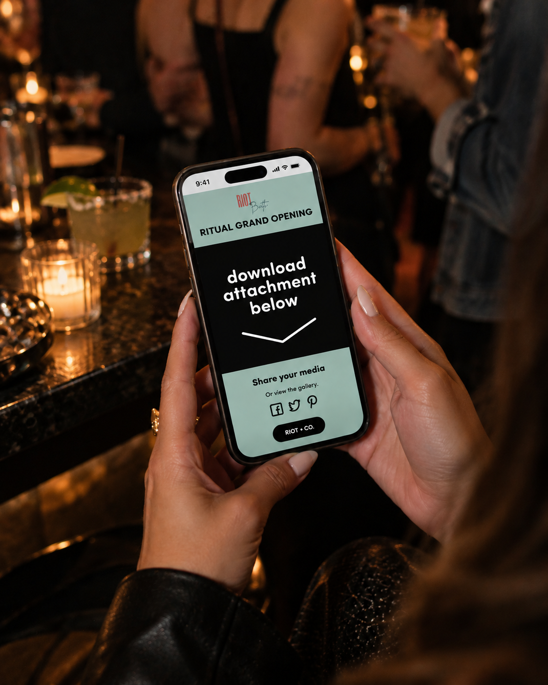 A person holding a smartphone displaying a digital flyer for the grand opening of a venue called Riot. The flyer instructs to download an attachment and share on social media platforms. The background features a dimly-lit bar with drinks, candles, and people.