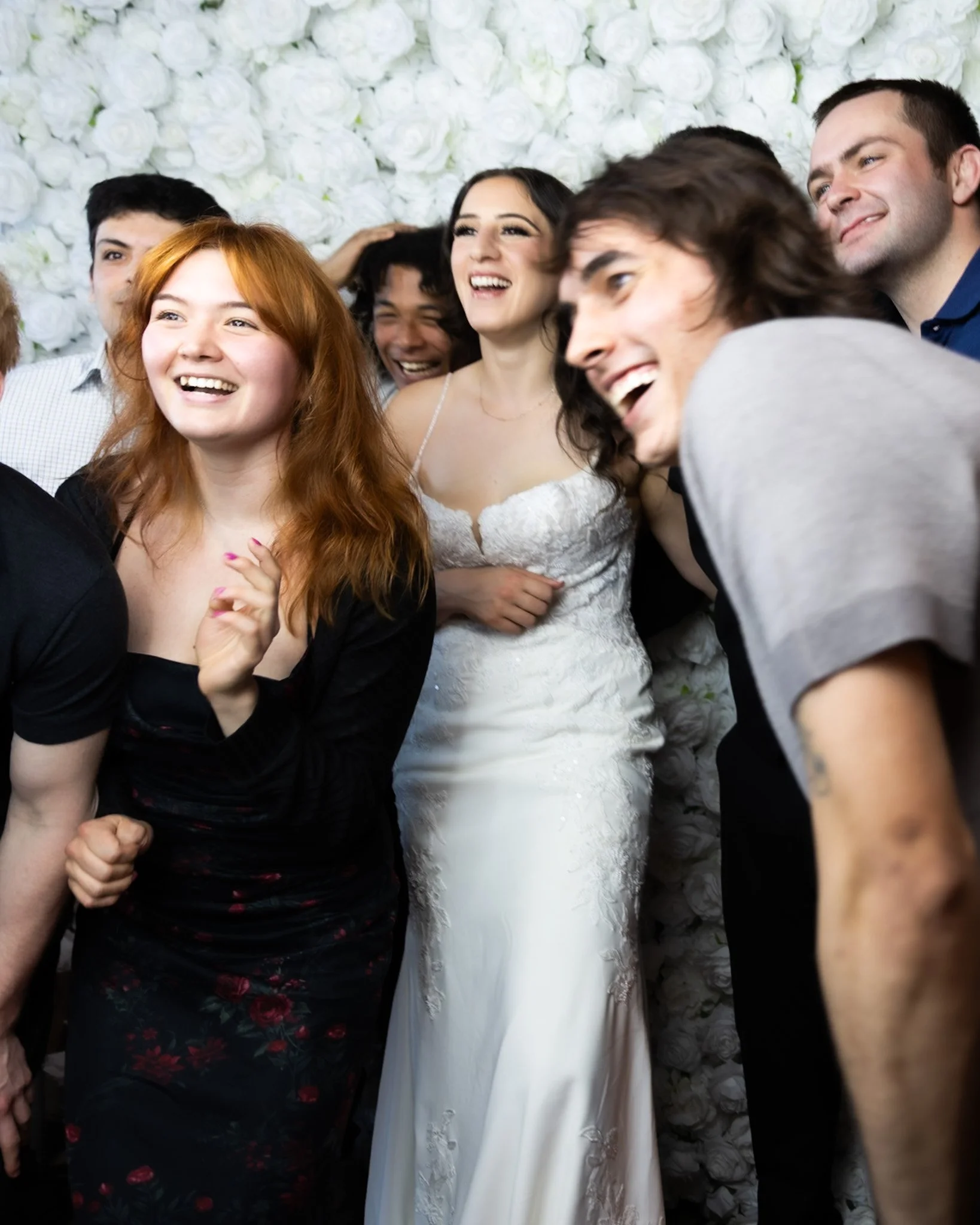 Group of people joyfully celebrating, with a woman in a white dress at the center, standing in front of a wall of white roses.