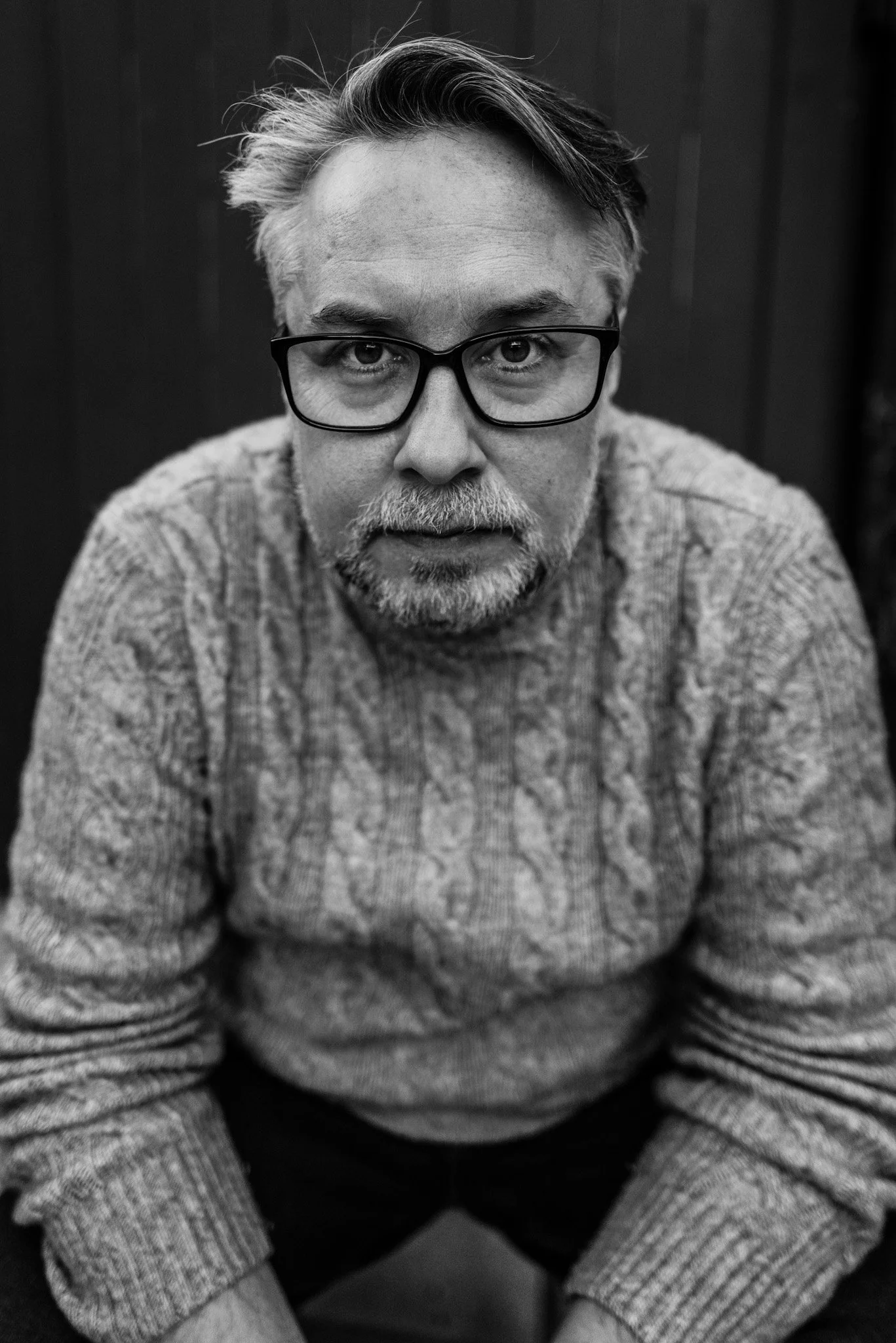 Black and white photo of a man with glasses and a beard, wearing a textured sweater, looking directly at the camera.