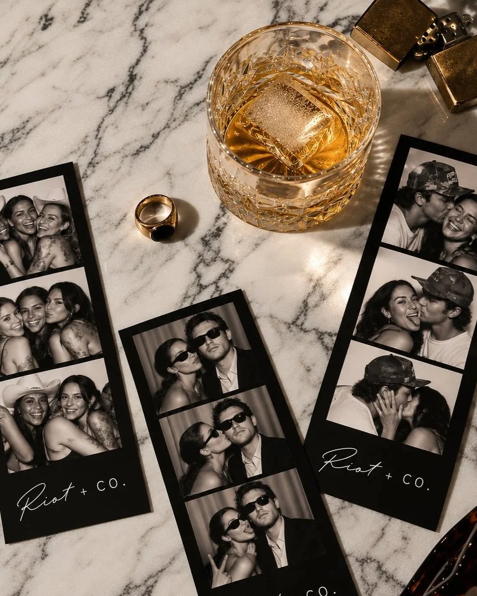 Photo booth strips with friends, a glass of whiskey with ice, a gold ring, and two gold lighters on a marble surface.