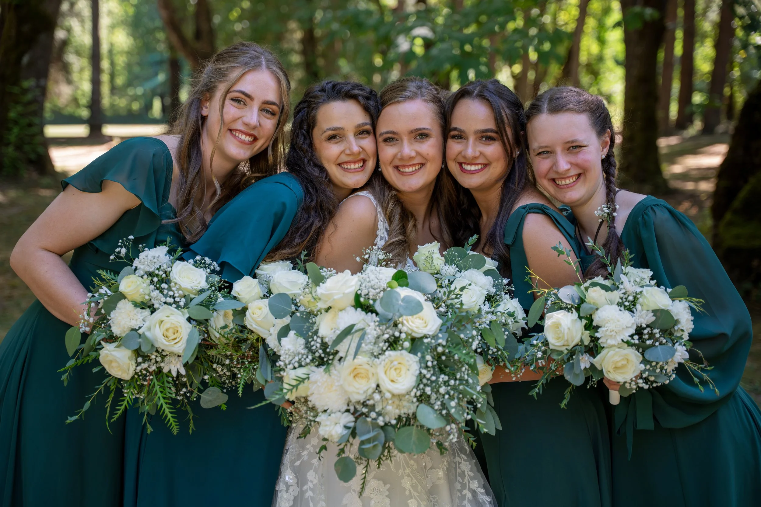 Group of five smiling women in teal dresses holding a large bouquet of white flowers, outdoors in a wooded area.