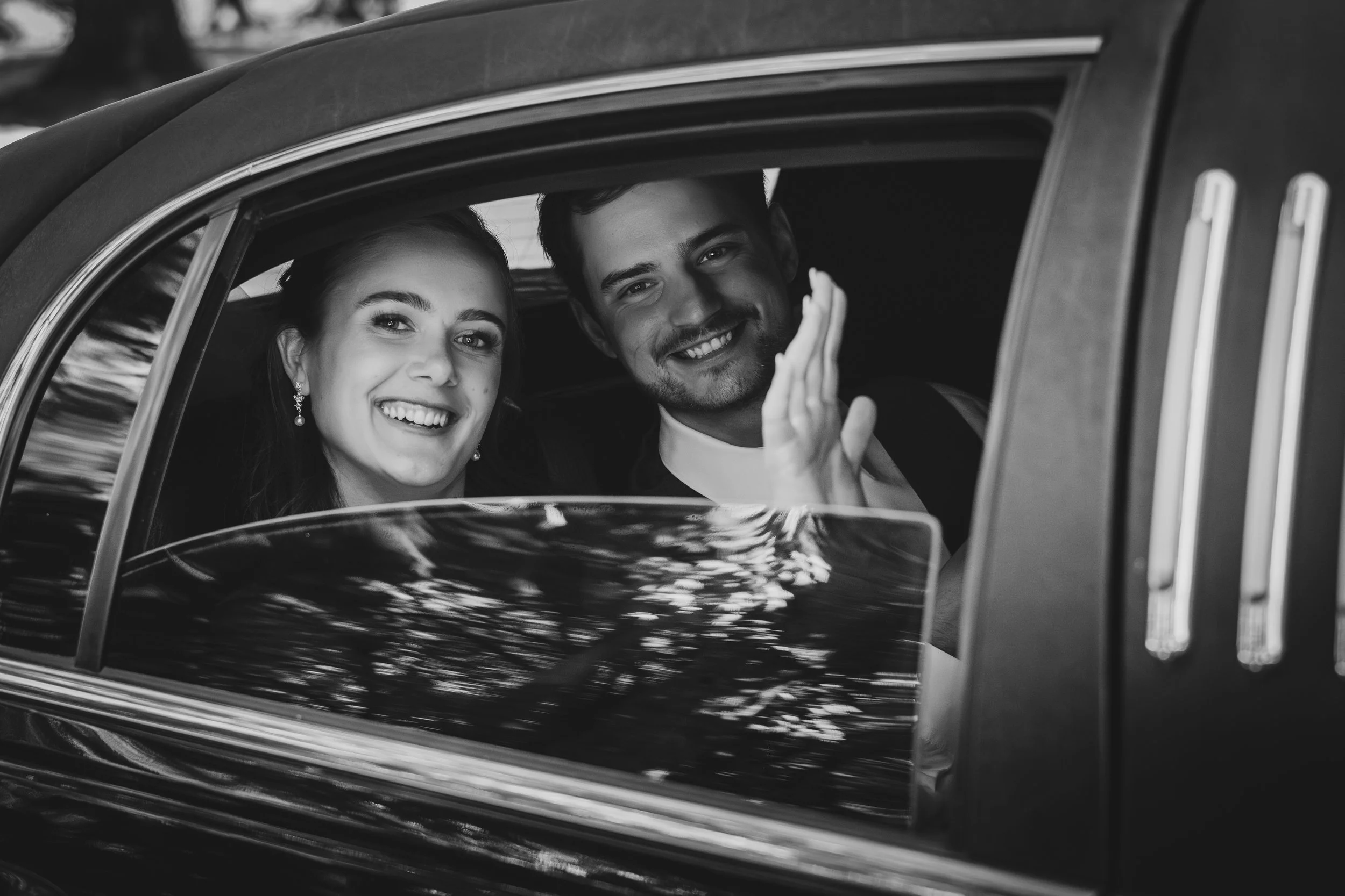 Black and white photo of a smiling woman with earrings and a smiling man waving as they sit inside a vintage car.