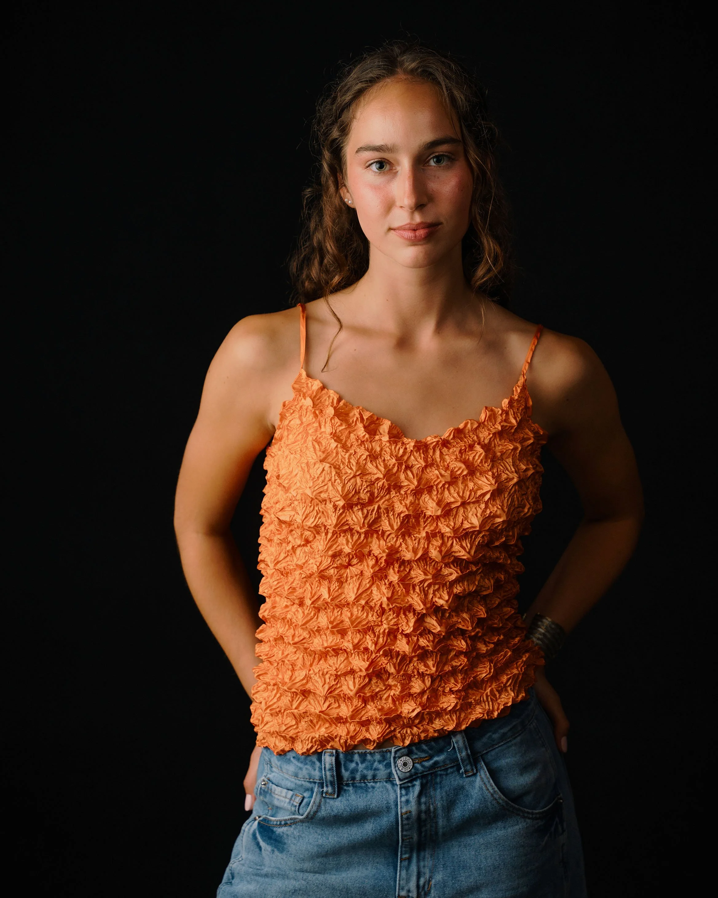 A young woman wearing a textured orange sleeveless top and jeans, standing against a black background.