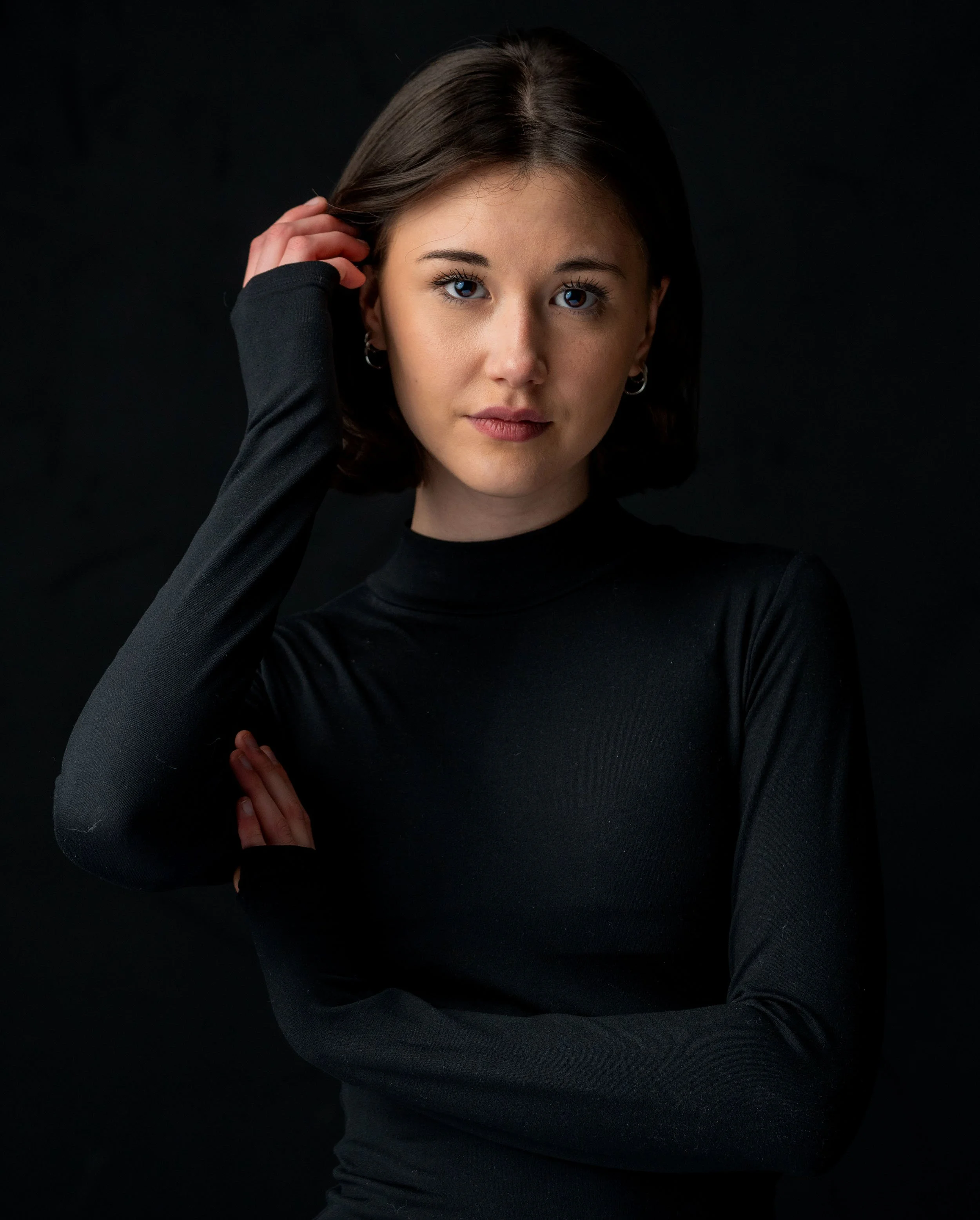 Young woman with dark hair and blue eyes, wearing a black long-sleeve top, posing against a black background.