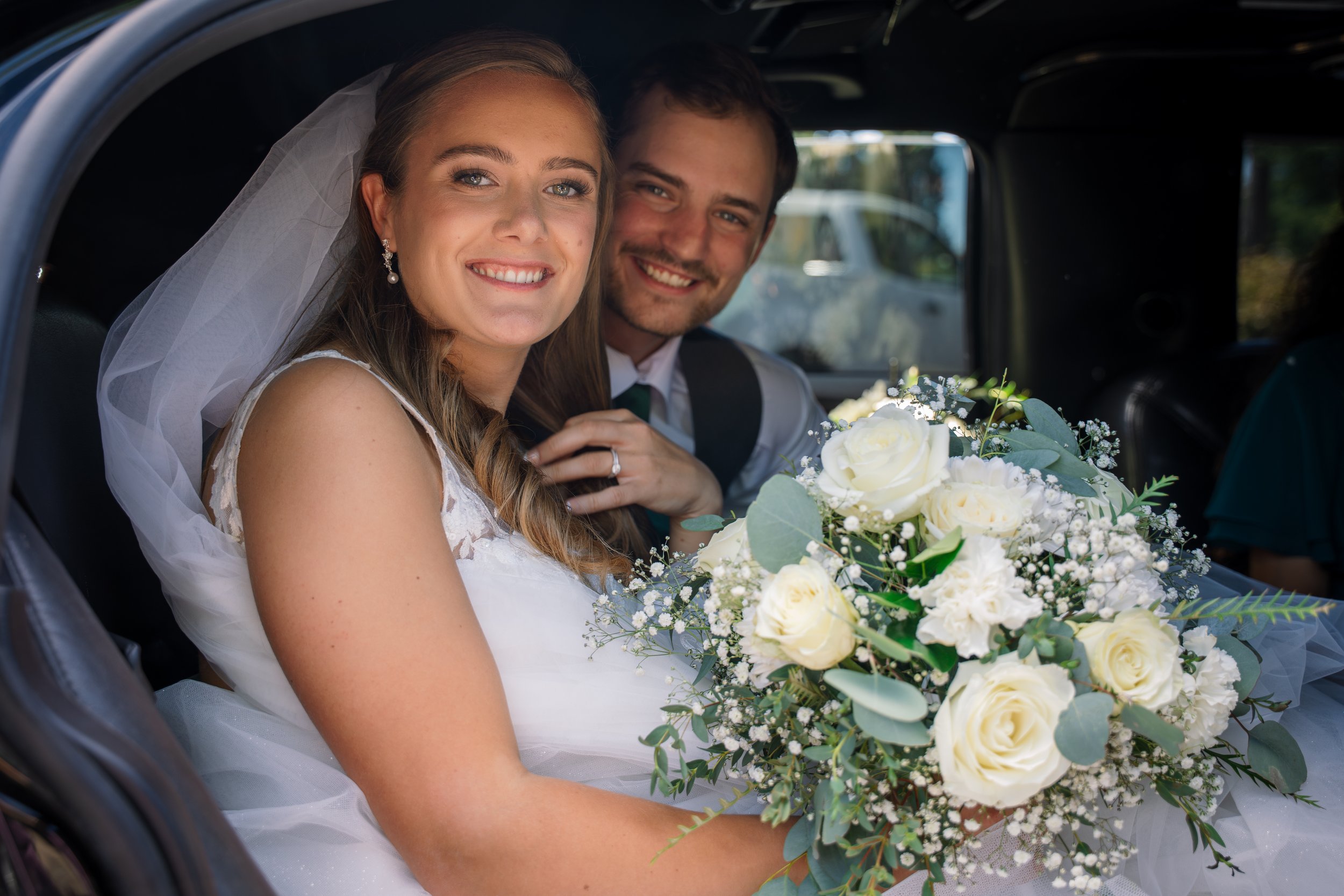 A smiling bride with a veil and earrings holding a bridal bouquet of white roses, sitting in a car during her wedding.