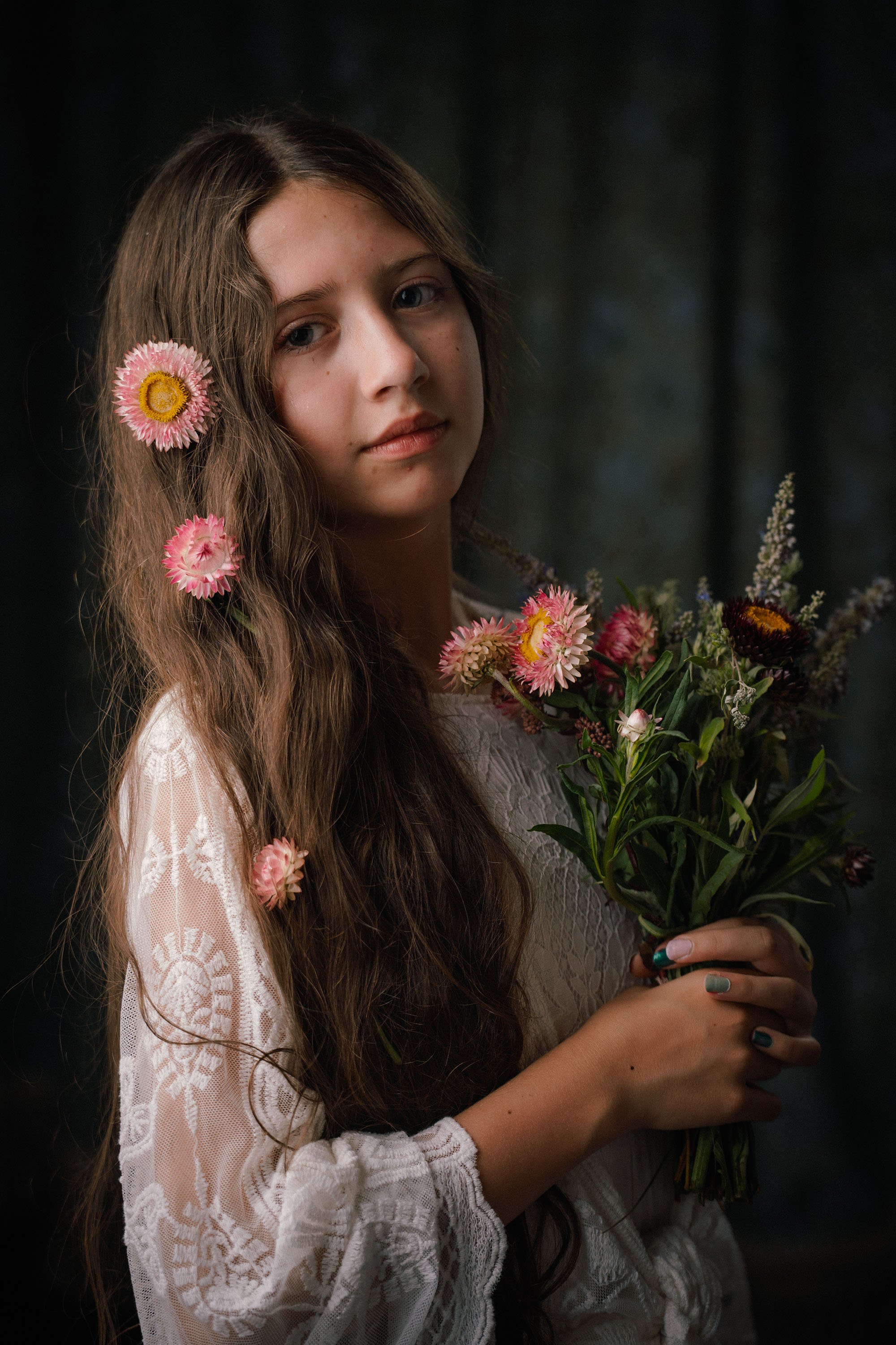 A young woman with long, wavy brown hair holding a bouquet of colorful flowers, wearing a white lace dress, and standing against a dark, blurred background.