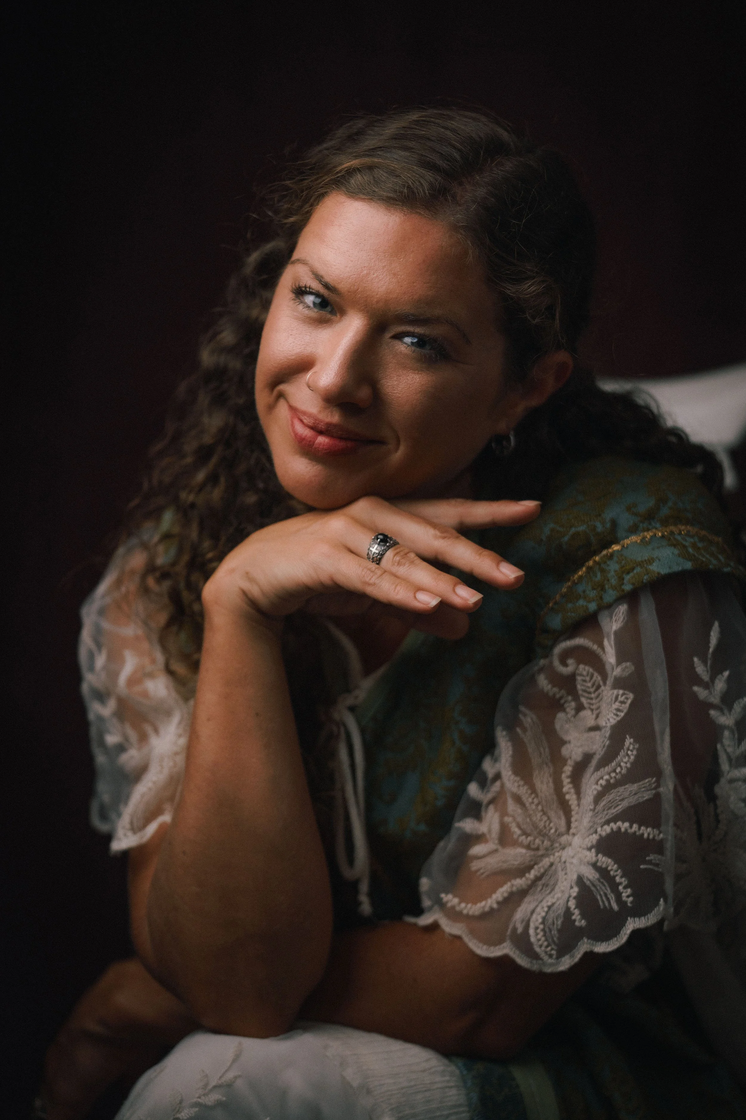 A woman with curly hair and a nose ring resting her chin on her hand, wearing a floral-patterned blouse, smiling softly, against a dark background.