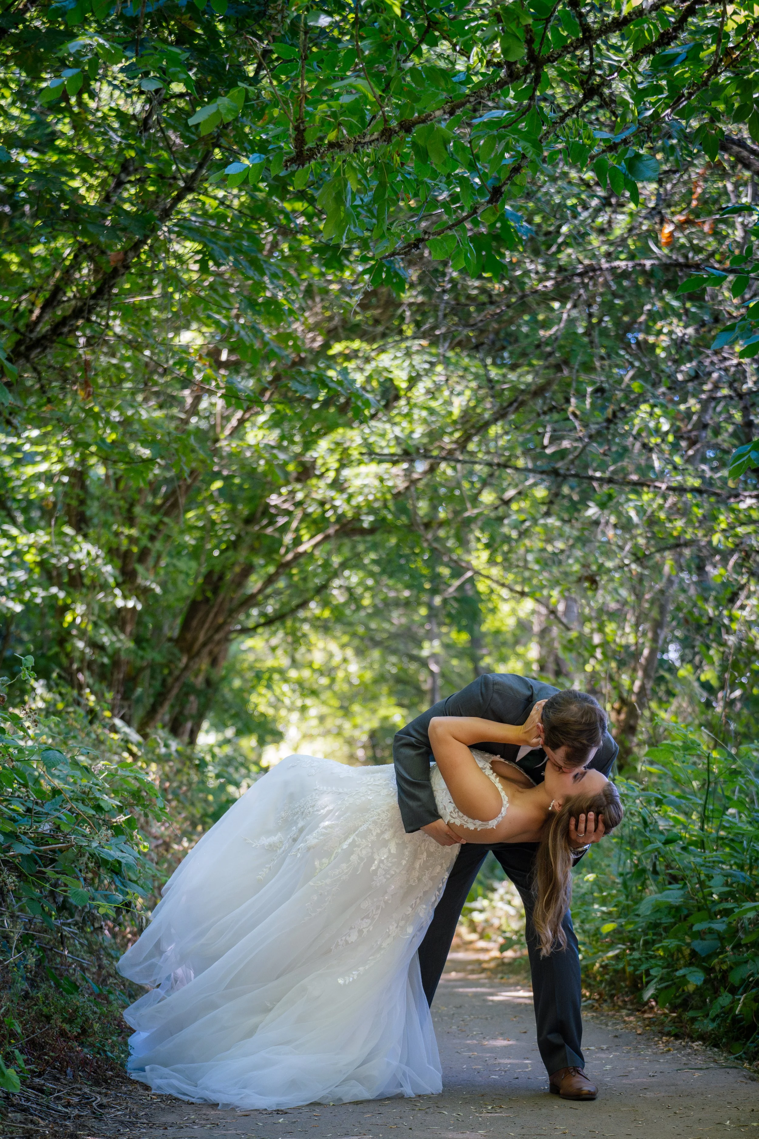 A bride and groom are sharing a kiss outdoors on a forest trail, with lush green trees overhead. The groom is lifting the bride as they kiss, and she is wearing a white wedding gown while he is dressed in a dark suit.