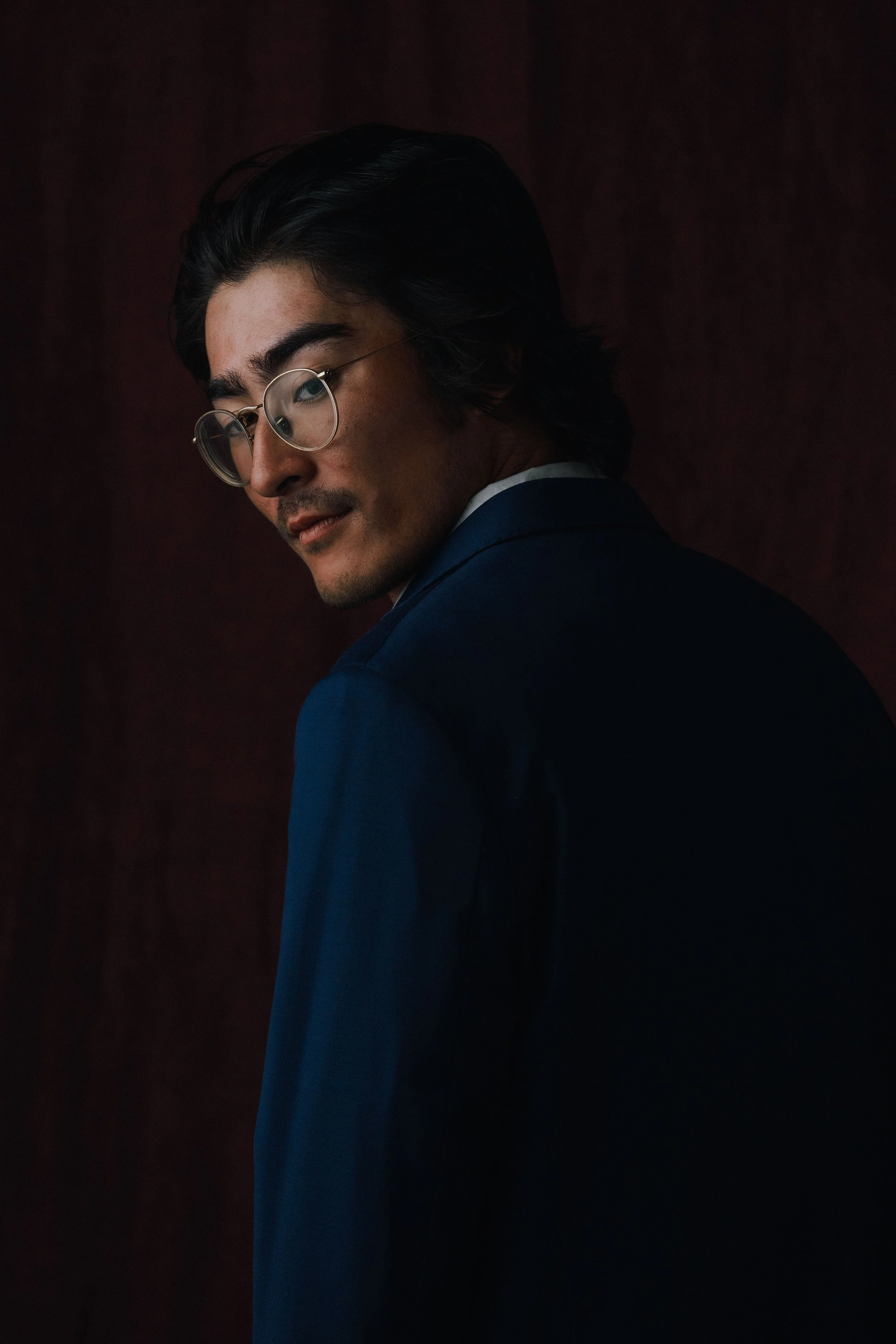 Close-up of a young man with black hair, glasses, and a blue suit, looking slightly downward with a subtle smile, against a dark, textured background.