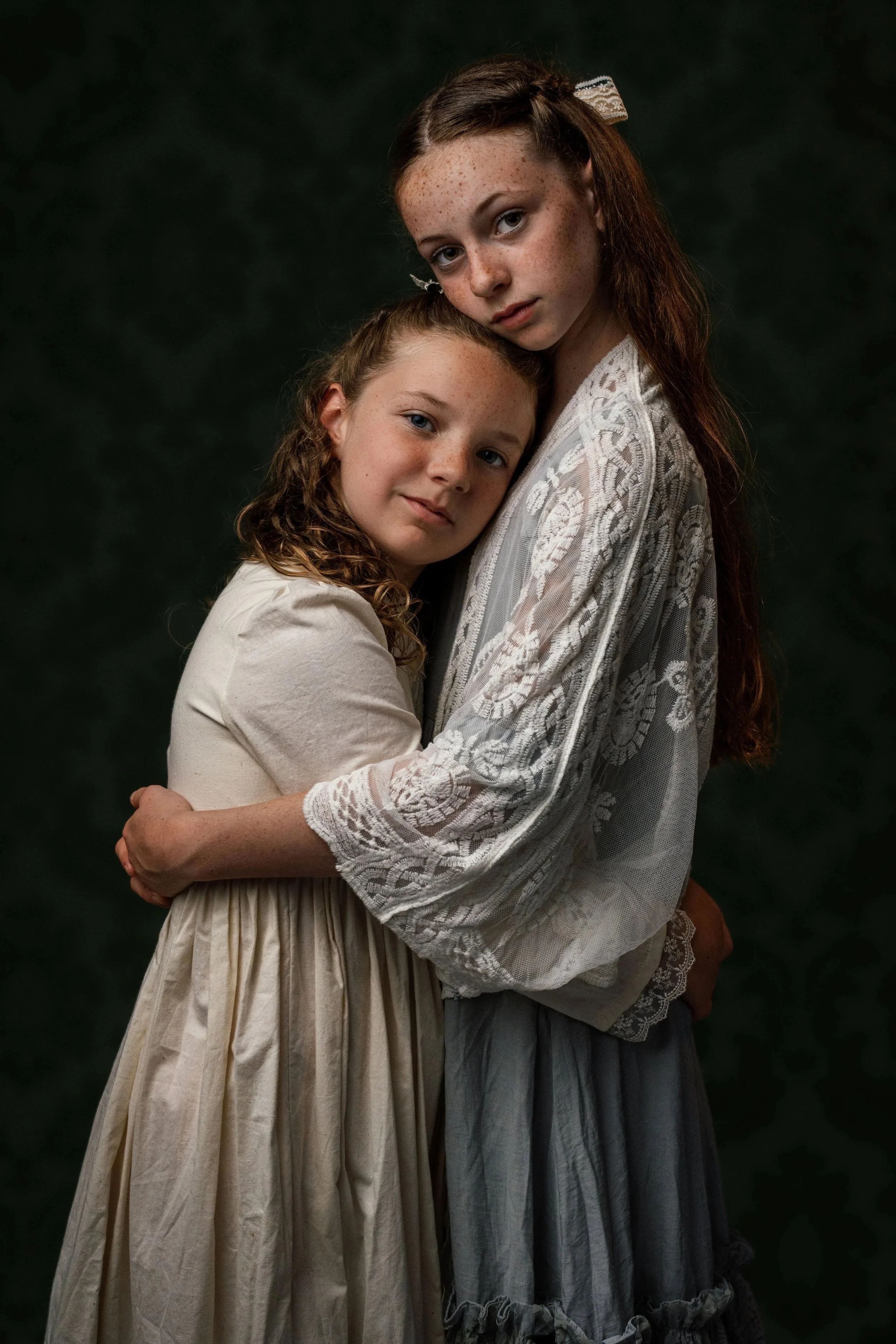 Two young girls hugging each other in front of a dark background. The girl on the right has long red hair, freckles, and is wearing a white lace top. The girl on the left has curly hair and is wearing a light-colored dress.