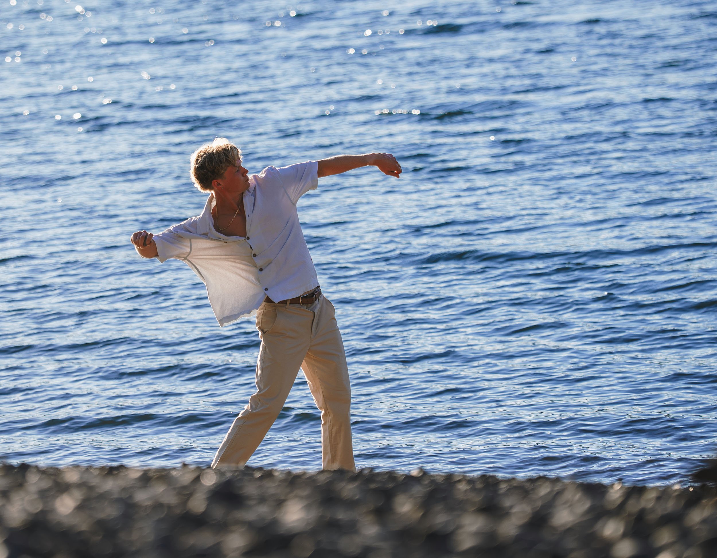A young man with blond hair, wearing a white shirt and beige pants, is standing along the shoreline, throwing a punch with his right fist towards the water, during daytime.