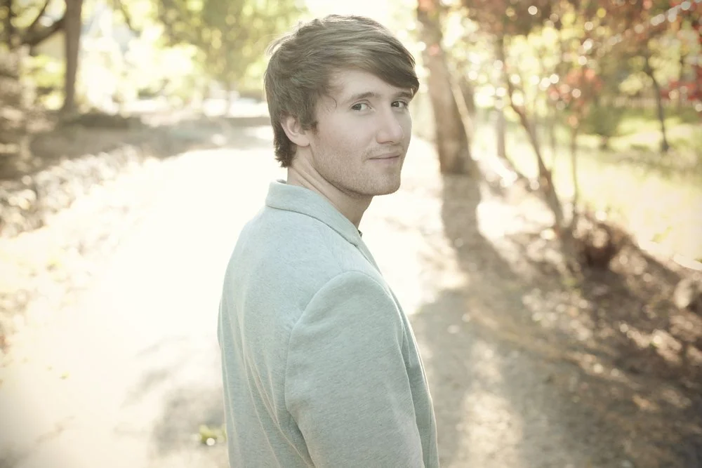 A young man with brown hair and a light gray hoodie standing outdoors on a sunlit path with autumn trees in the background.