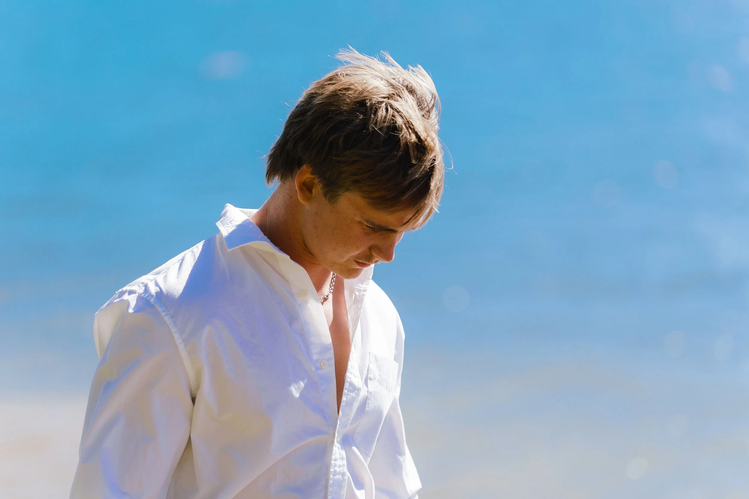 A young man with light brown hair and fair skin looking downwards in a bright outdoor setting with a clear blue sky.