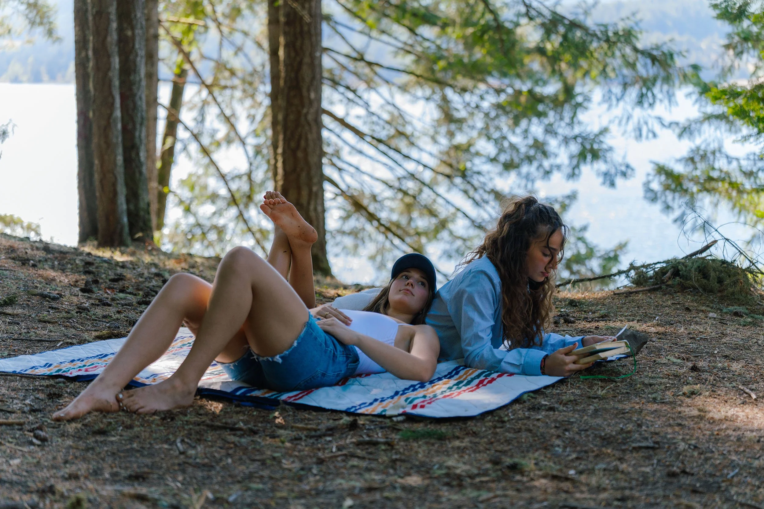 Two young women and a young girl lie on a blanket in a forested area by a lake, surrounded by tall trees with green foliage, under a blue sky with clouds.