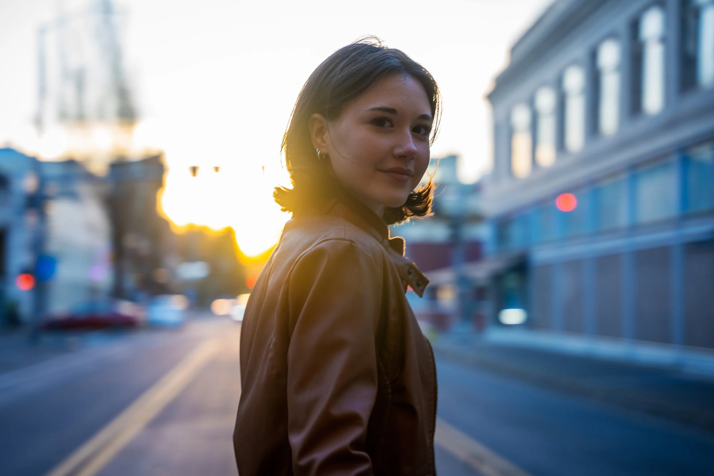 A woman with shoulder-length dark hair and earrings, wearing a brown jacket, standing outdoors in a city at sunset with blurred buildings and cars in the background.