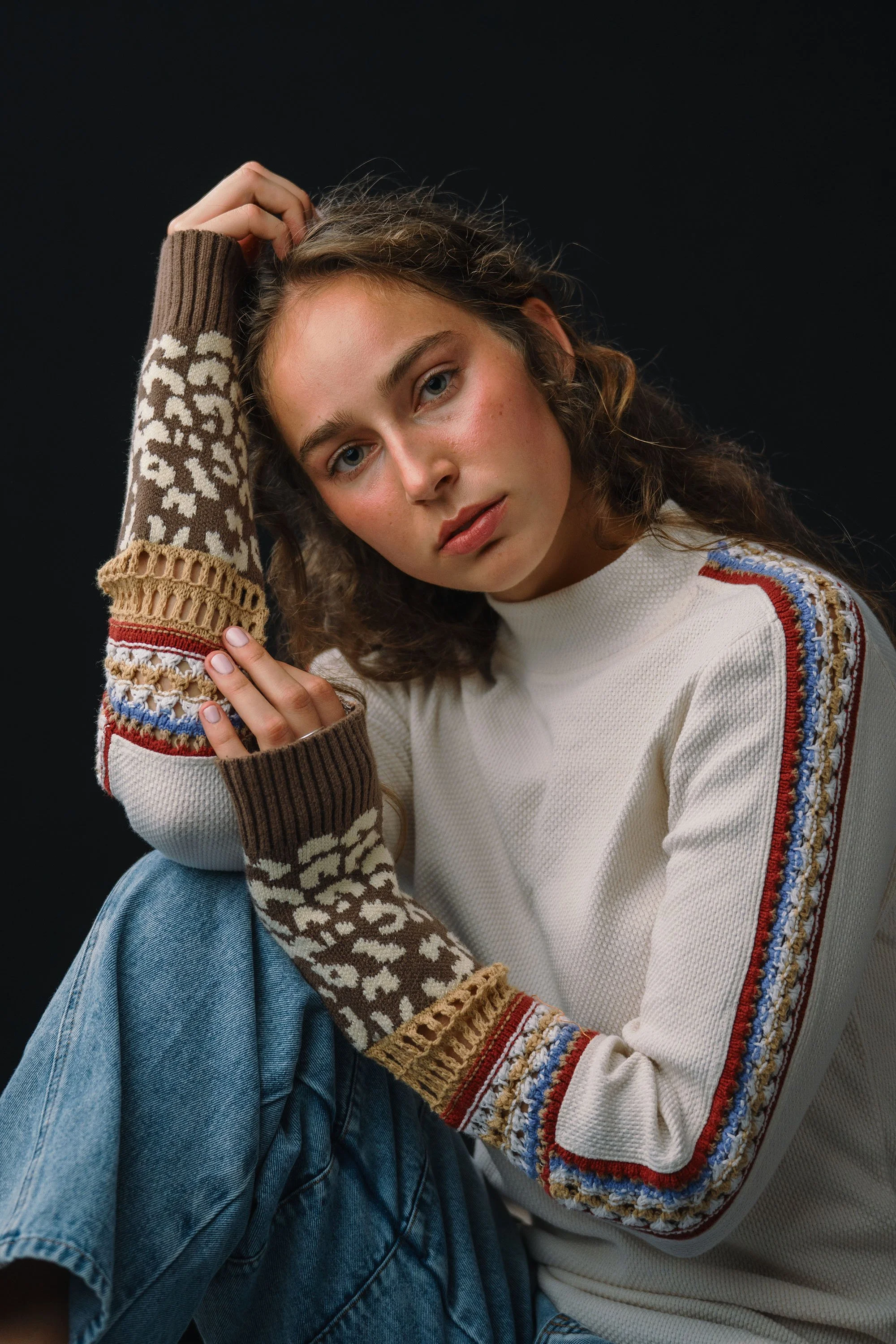 A young woman with curly hair and fair skin posing against a black background, wearing a white sweater with colorful embroidery on the sleeves and blue jeans.