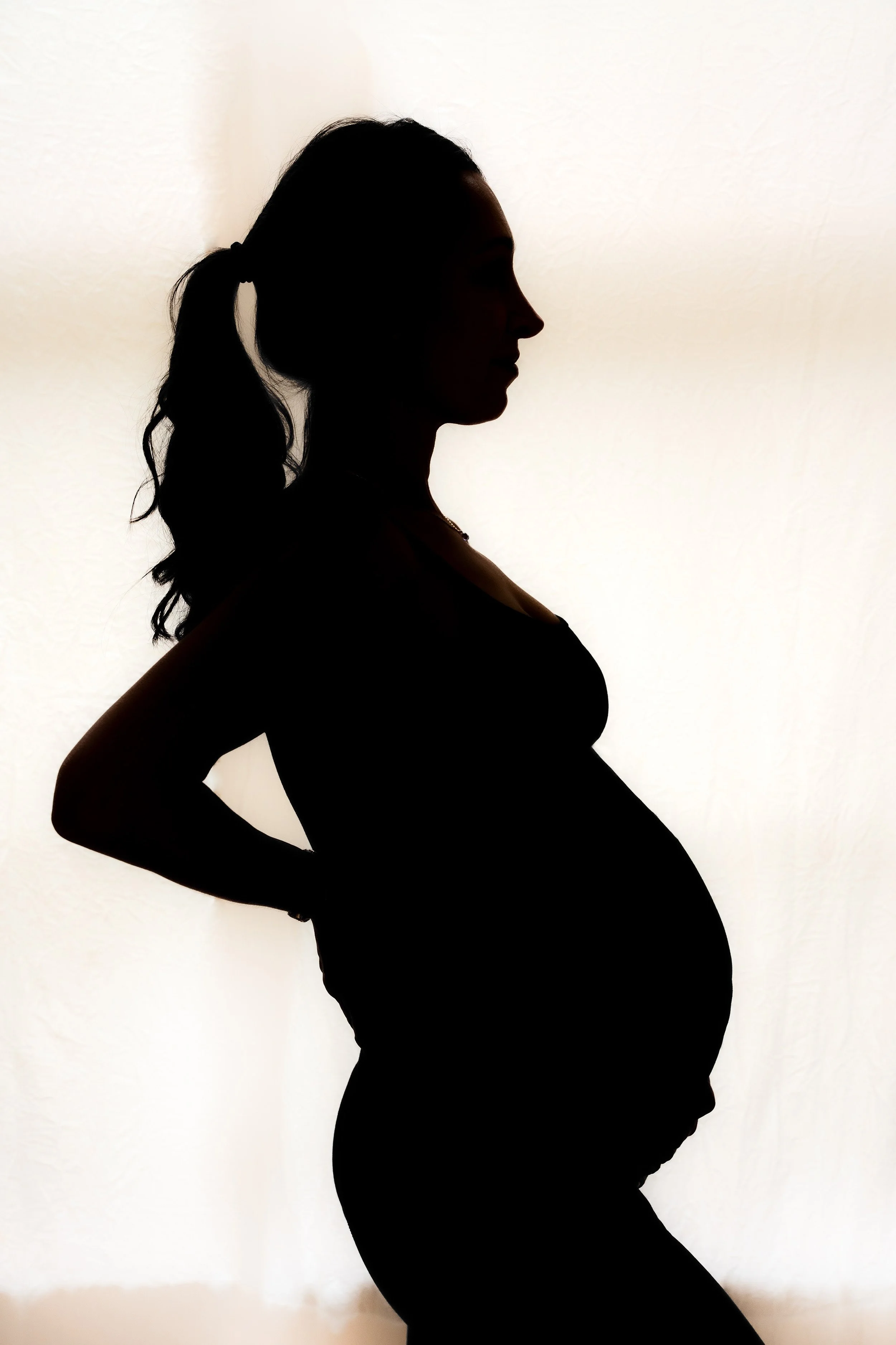 Silhouette of a pregnant woman with her hands on her hips, standing sideways against a plain background.
