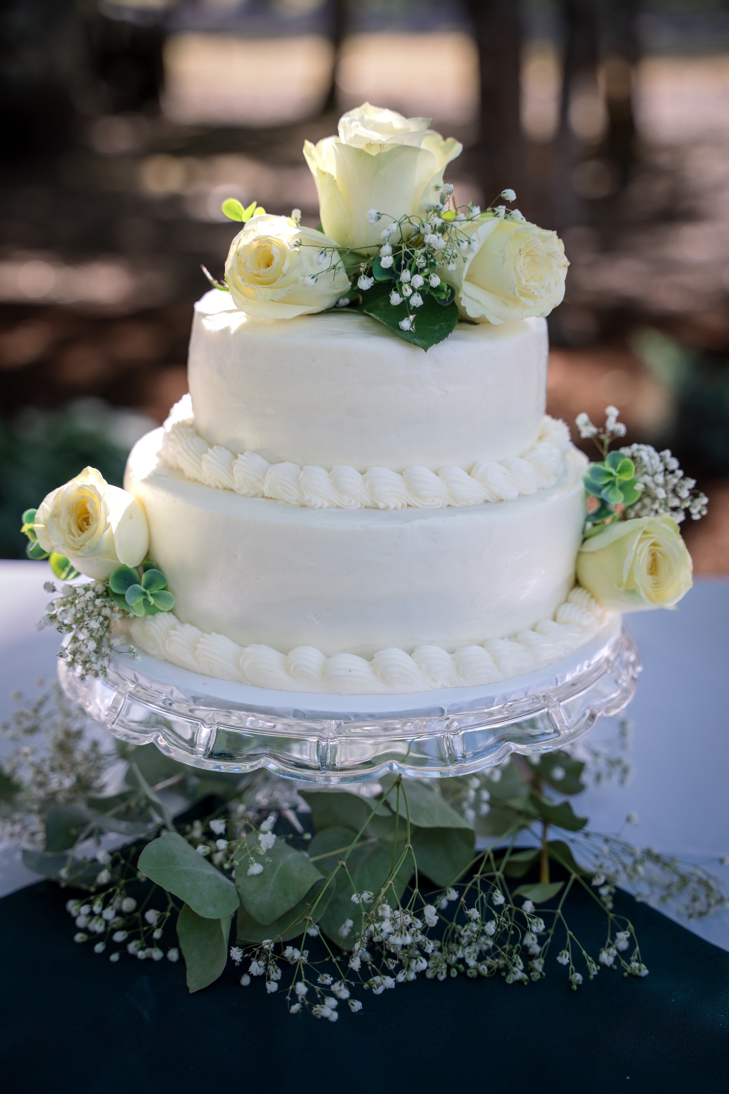 A white wedding cake decorated with white roses, baby's breath, and greenery, placed on a glass cake stand with a dark cloth underneath and a blurred outdoor background.