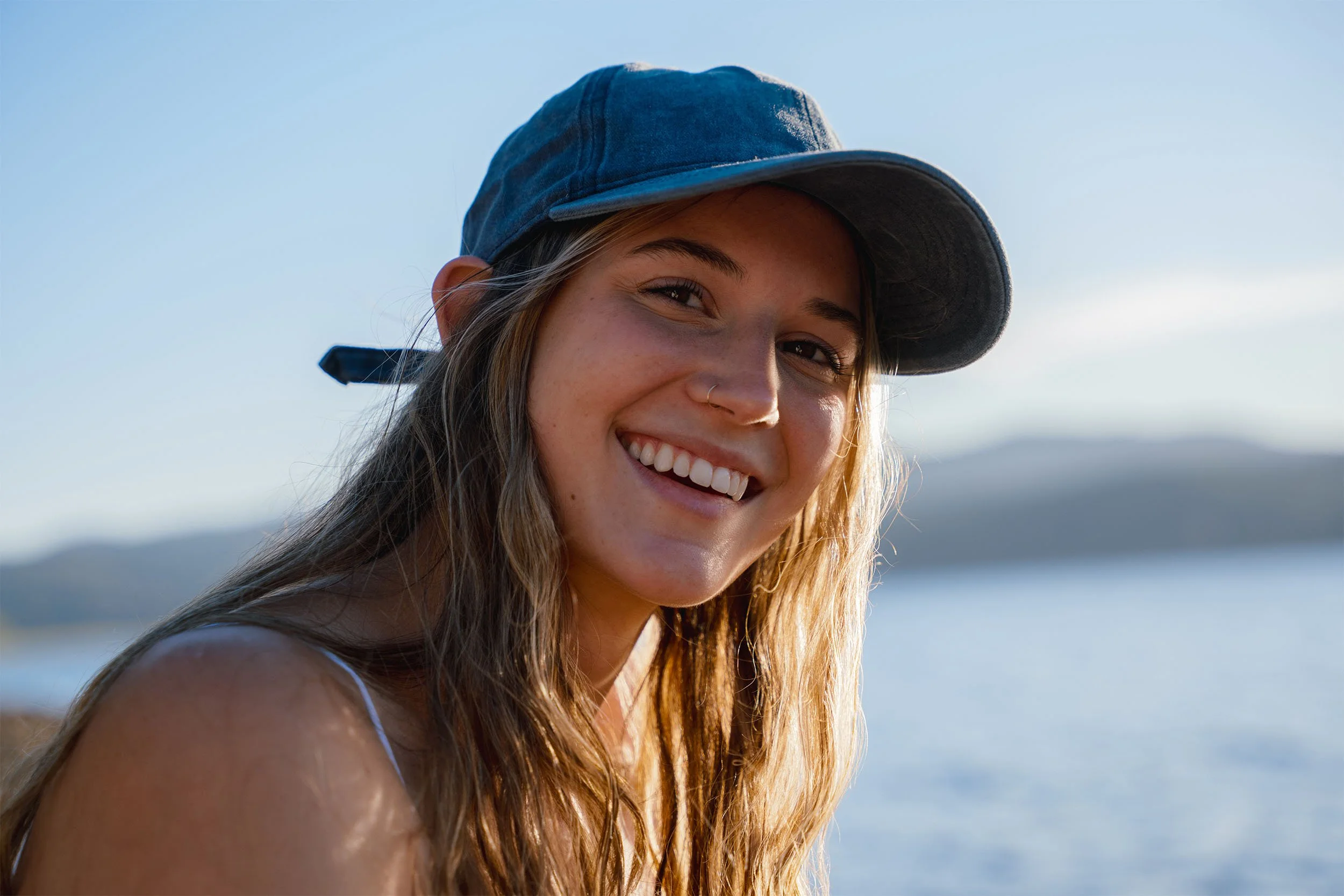 A young woman with long, wavy hair wearing a denim cap smiling outdoors near water during daytime.
