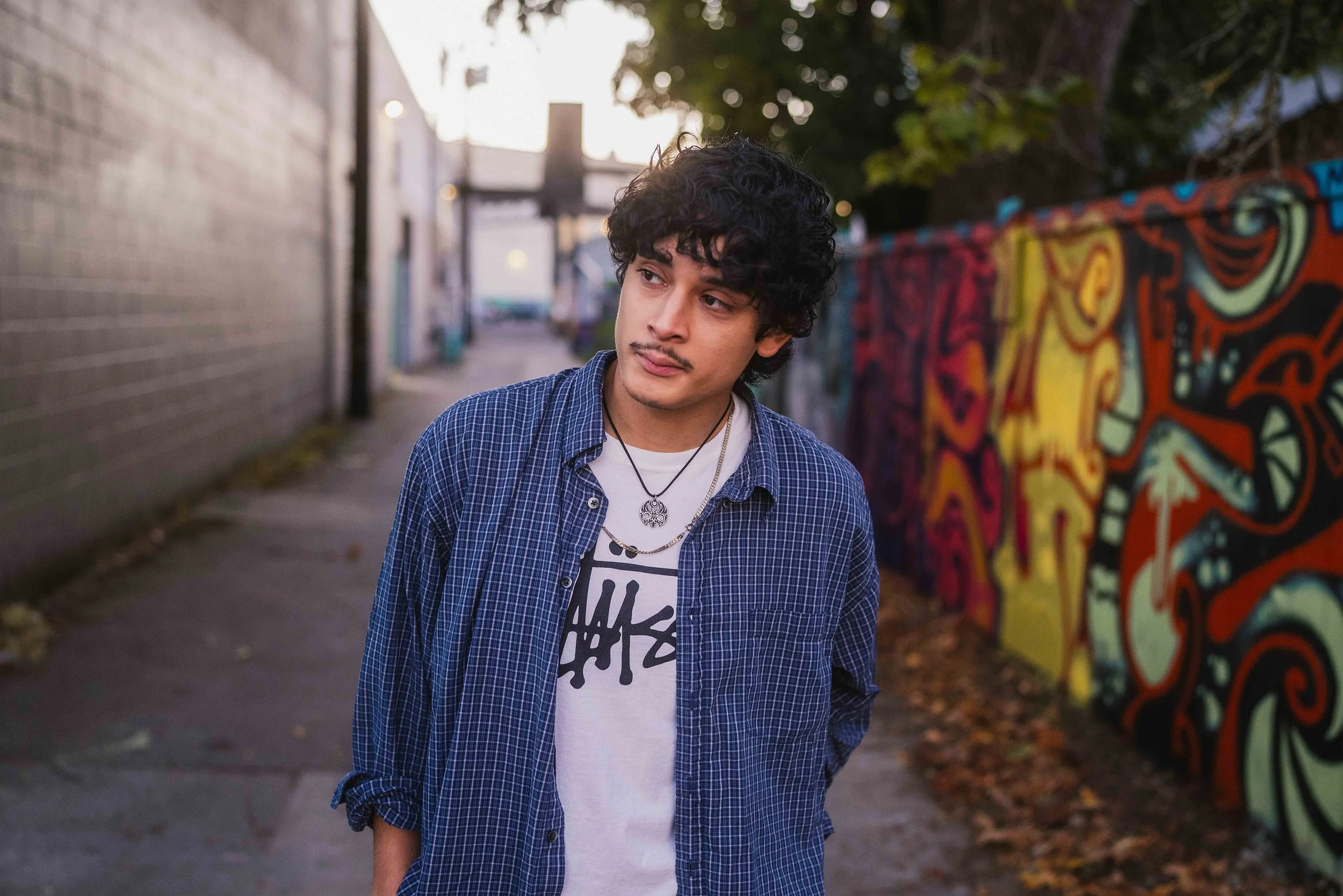 Young man with curly dark hair and a mustache, wearing a blue plaid shirt over a graphic T-shirt and necklaces, standing outdoors near a colorful graffiti wall during daytime.