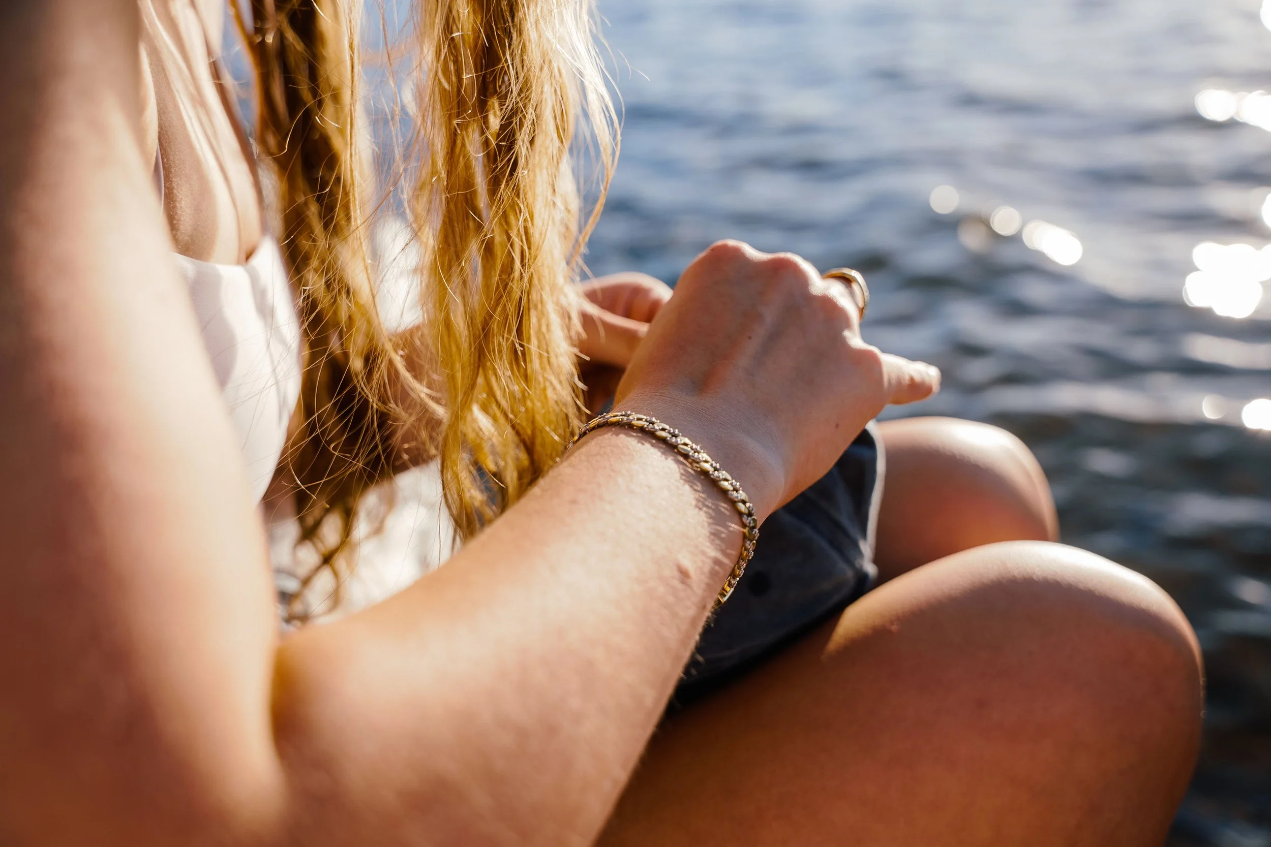 Close-up of a woman with long red hair sitting near a body of water, with sunlight reflecting on the water's surface.