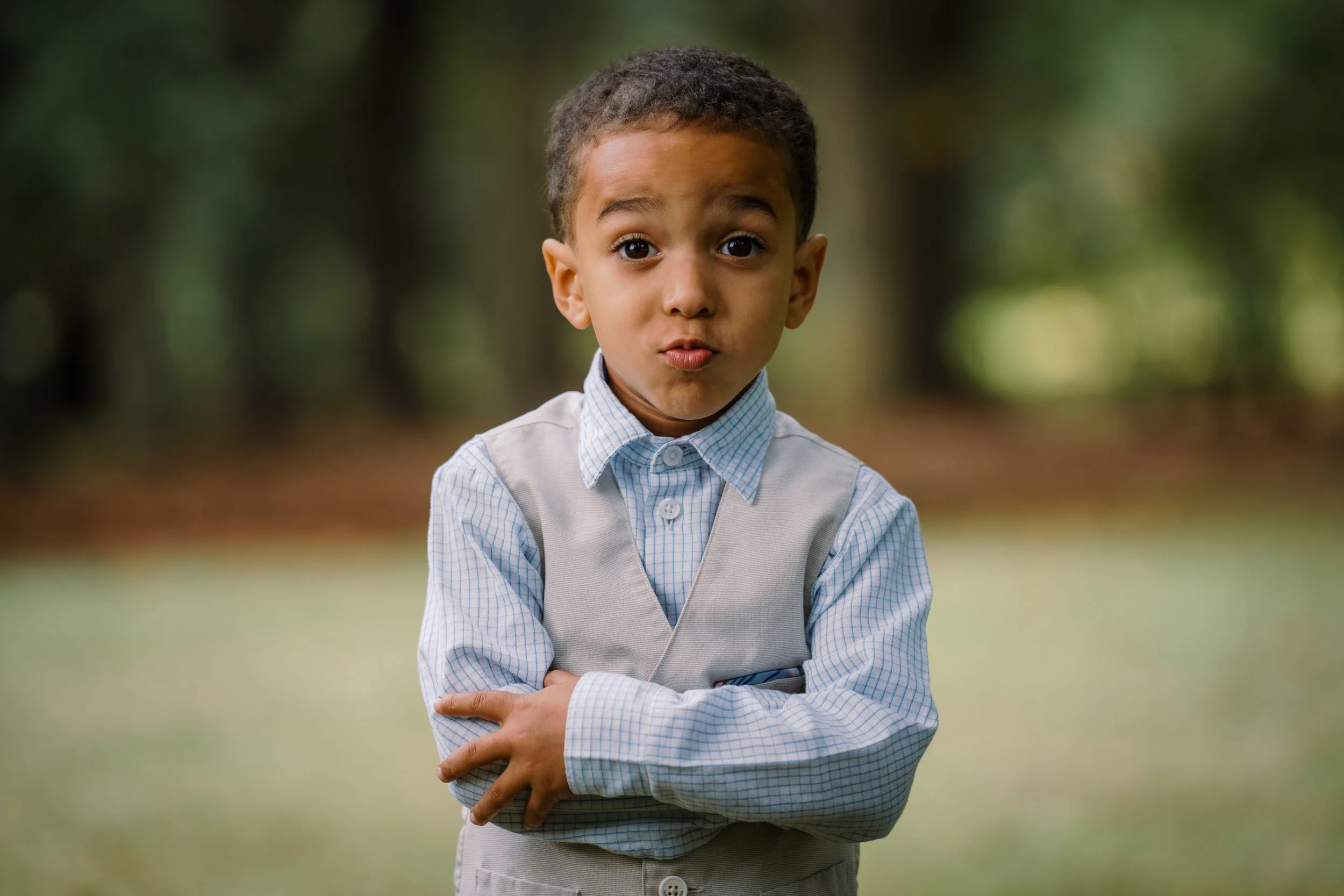 A young boy with curly hair making a surprised face with his lips pursed and eyes wide open, standing outdoors with blurred trees in the background, wearing a light blue checkered shirt and a beige vest.