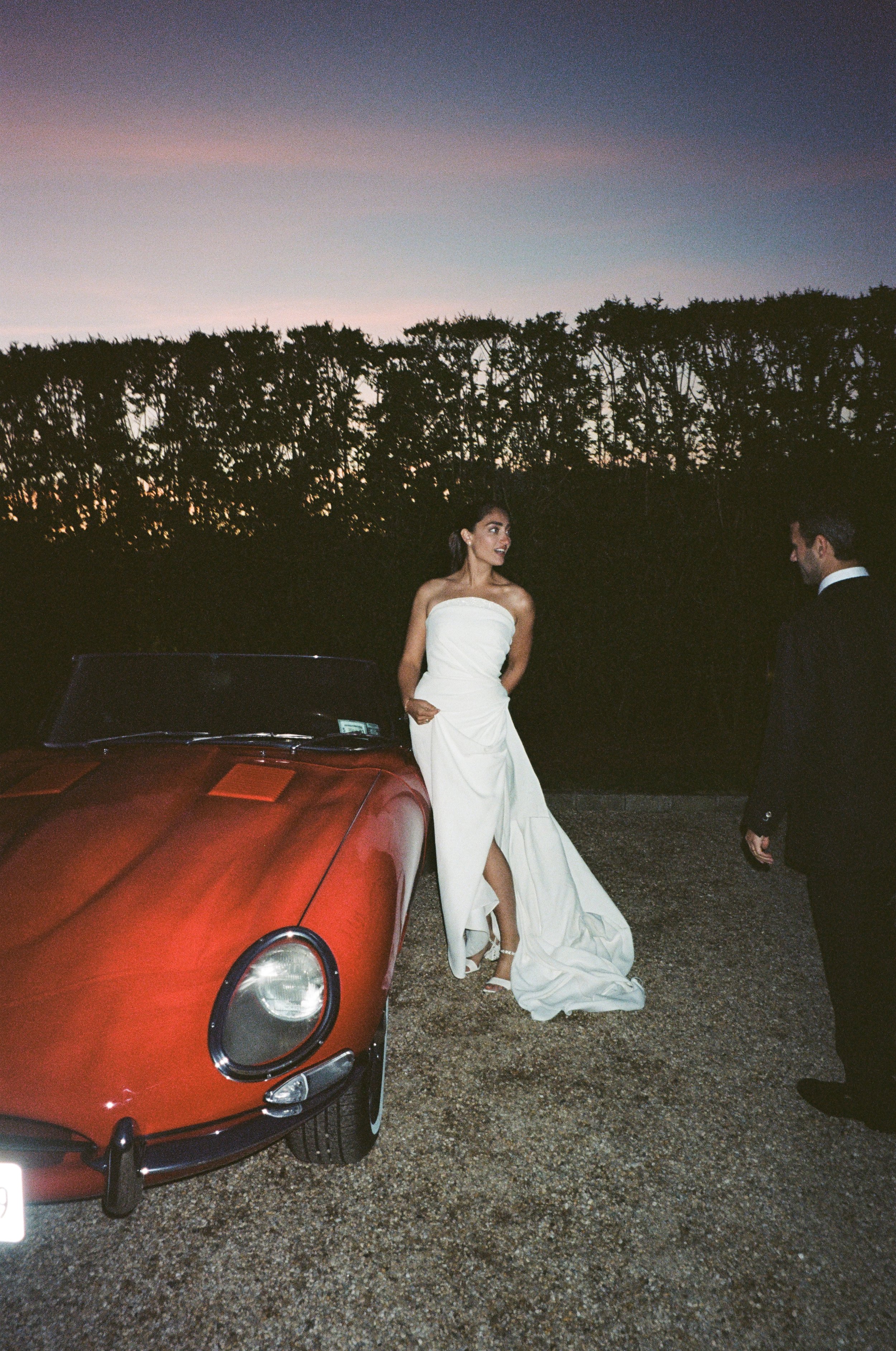 A woman in a strapless white wedding dress with a long train and a bride's bouquet is standing next to a red sports car at sunset. A man in a suit is standing nearby, and they are outdoors with a backdrop of trees and a colorful sky.