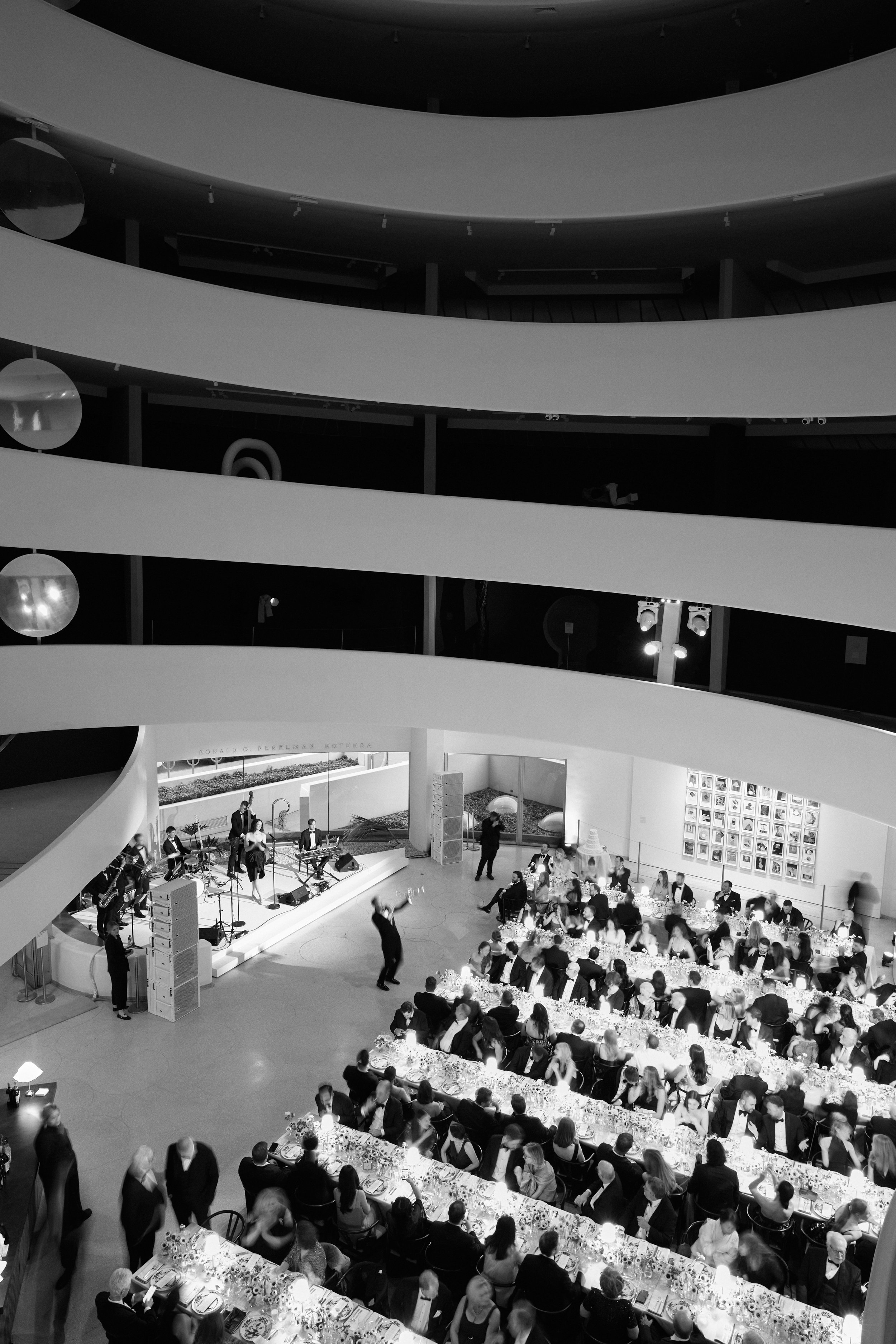 A large indoor banquet hall filled with guests seated at long tables decorated with floral centerpieces, and a musical performance happening on a stage. The space has multiple stories with curved balconies and hanging spherical lamps.