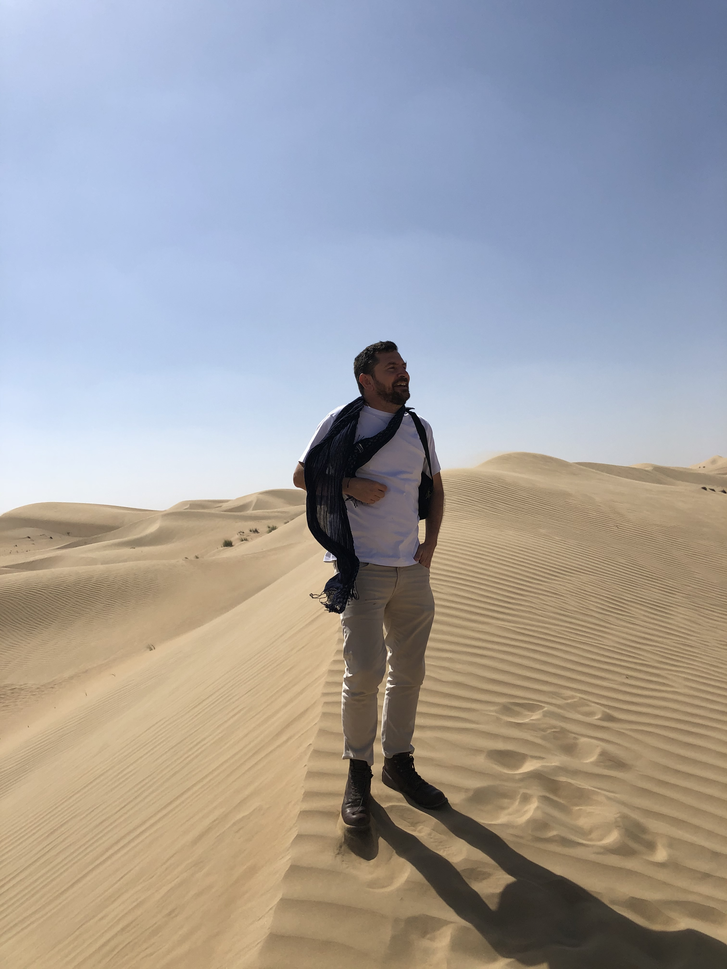 Man standing on sand dunes in a desert, wearing a white t-shirt, beige pants, black boots, and a black scarf suspension around his neck. The sky is clear and blue.