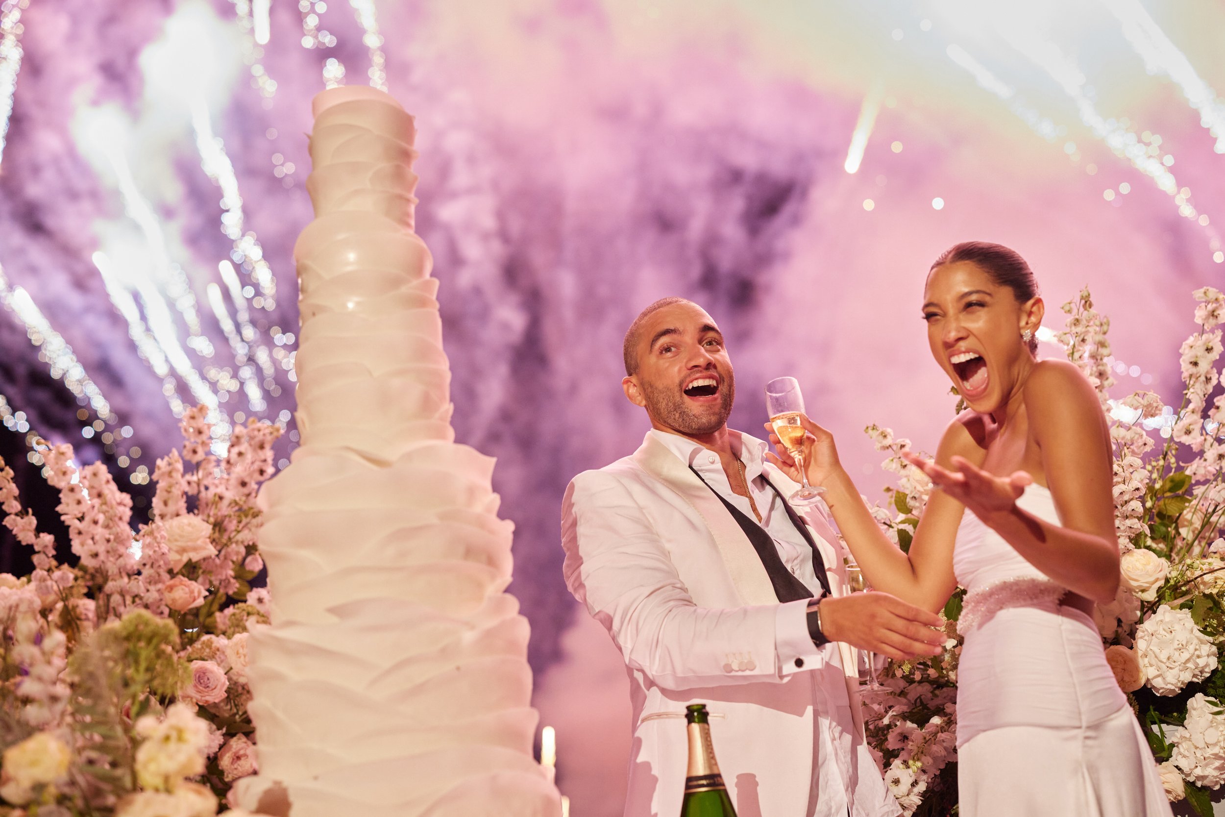 A happy couple celebrating at their wedding reception with champagne, surrounded by pink flowers, a tall wedding cake, fireworks in the background, and warm lighting.