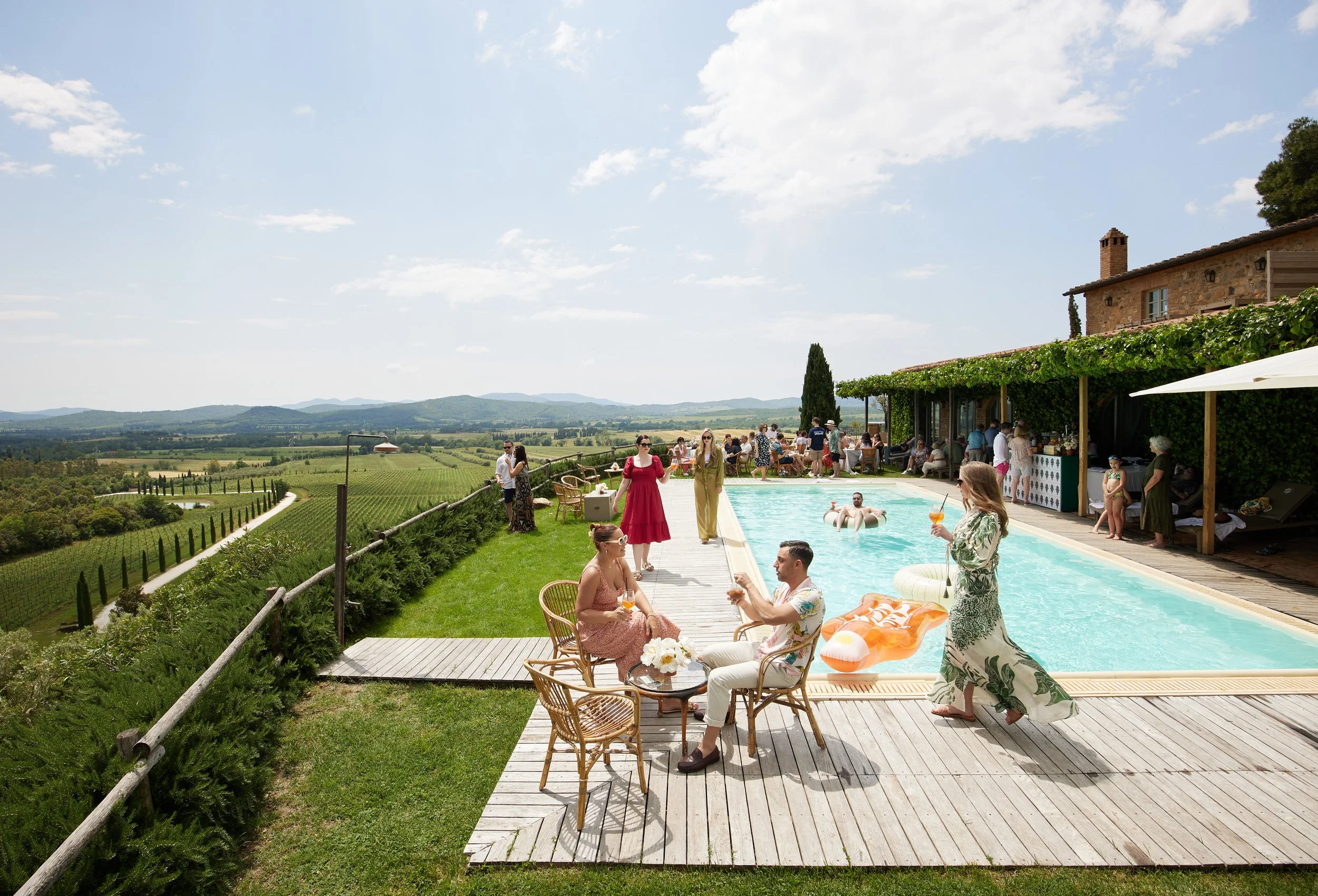 People enjoying a poolside gathering at a villa overlooking scenic vineyards and rolling hills under a partly cloudy sky.