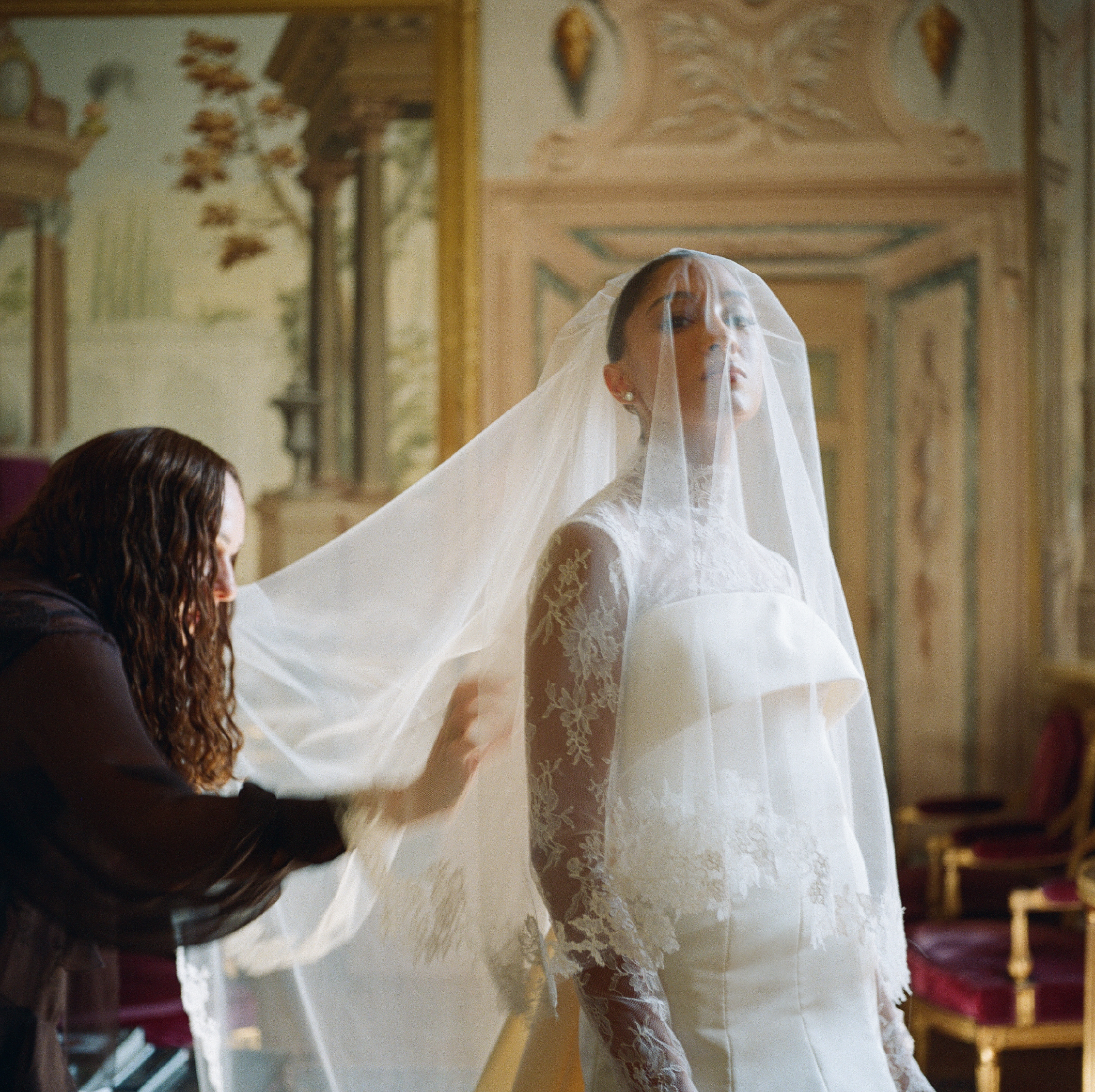A woman in a wedding dress wearing a veil, looking over her shoulder, in a richly decorated room with elaborate ceiling moldings and framed artwork.