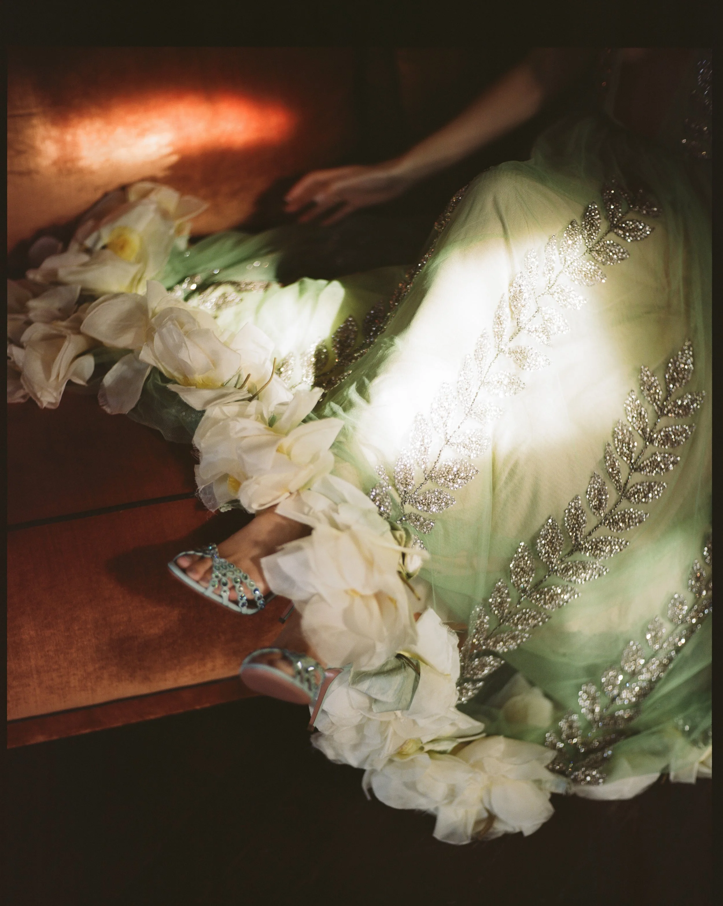 A woman wearing a light green, embellished sari sitting on a wooden surface with a bouquet of white flowers. The photo is taken at an angle with some shadowing and a dark background.