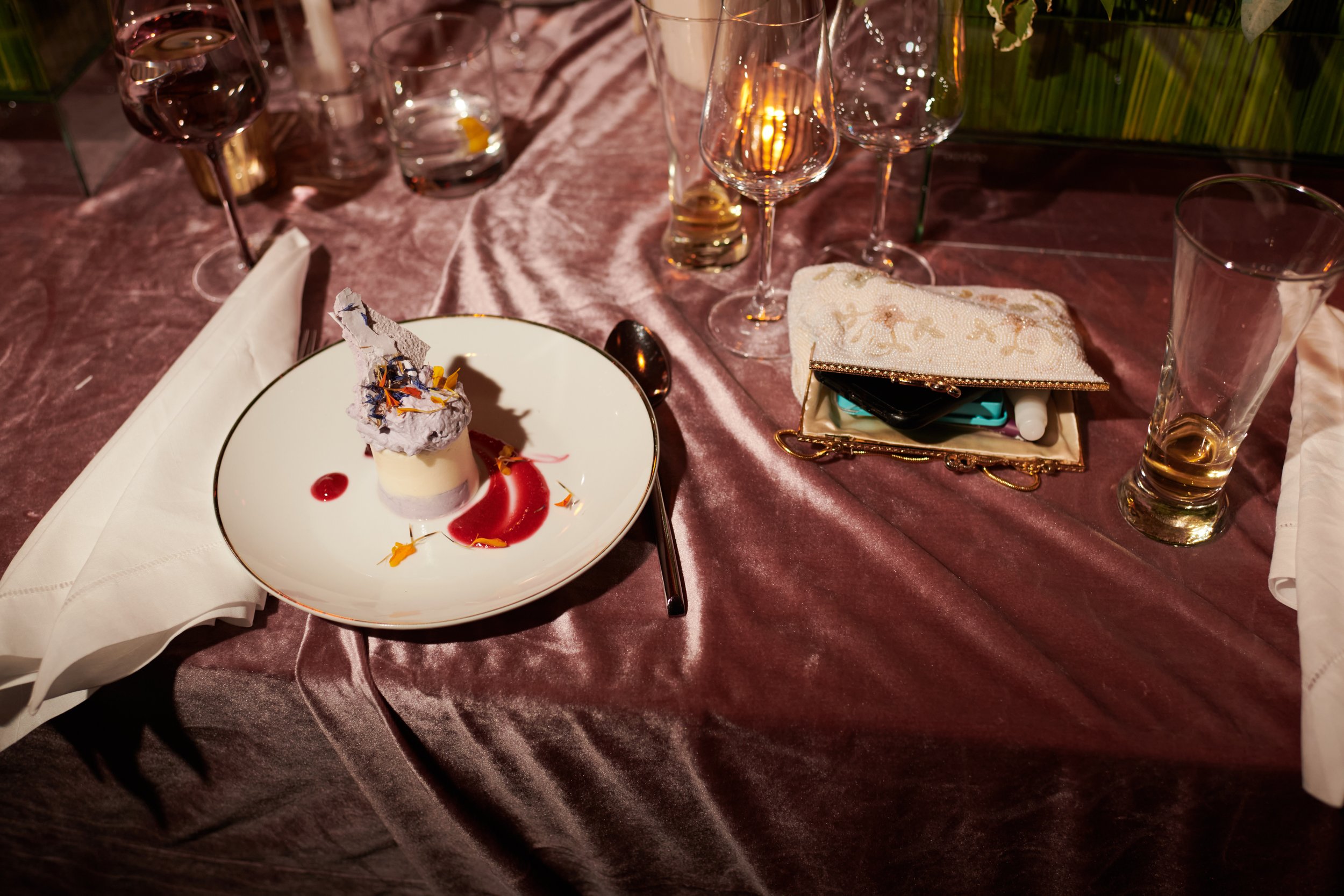 A dessert on a white plate, garnished with edible flowers and a decorative piece, accompanied by a red sauce. The table is set with various empty glasses, a napkin, and a gold-trimmed purse or clutch on a pink tablecloth.