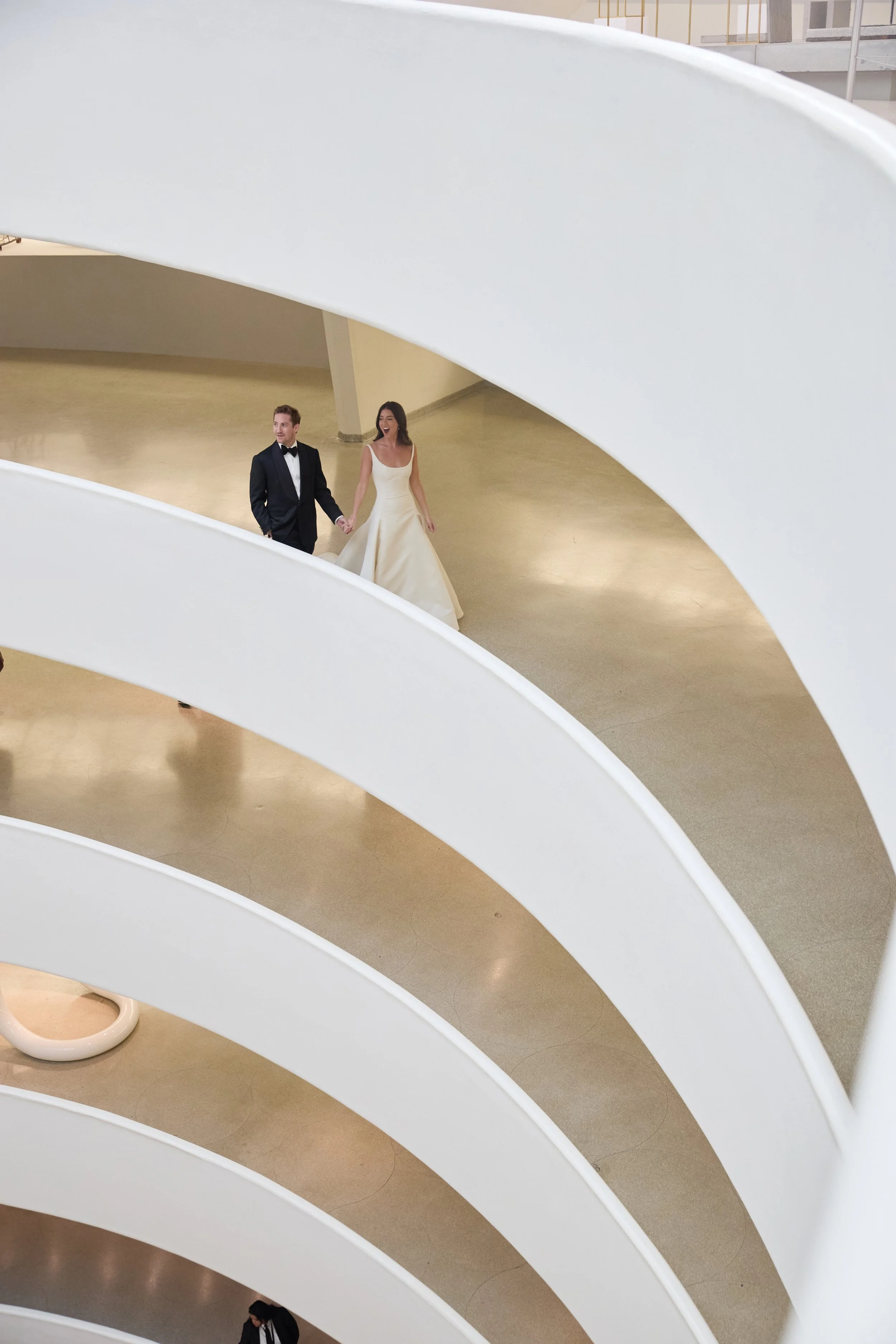 A couple dressed in formal wedding attire walking hand in hand inside a modern, white, curved building with multiple levels.
