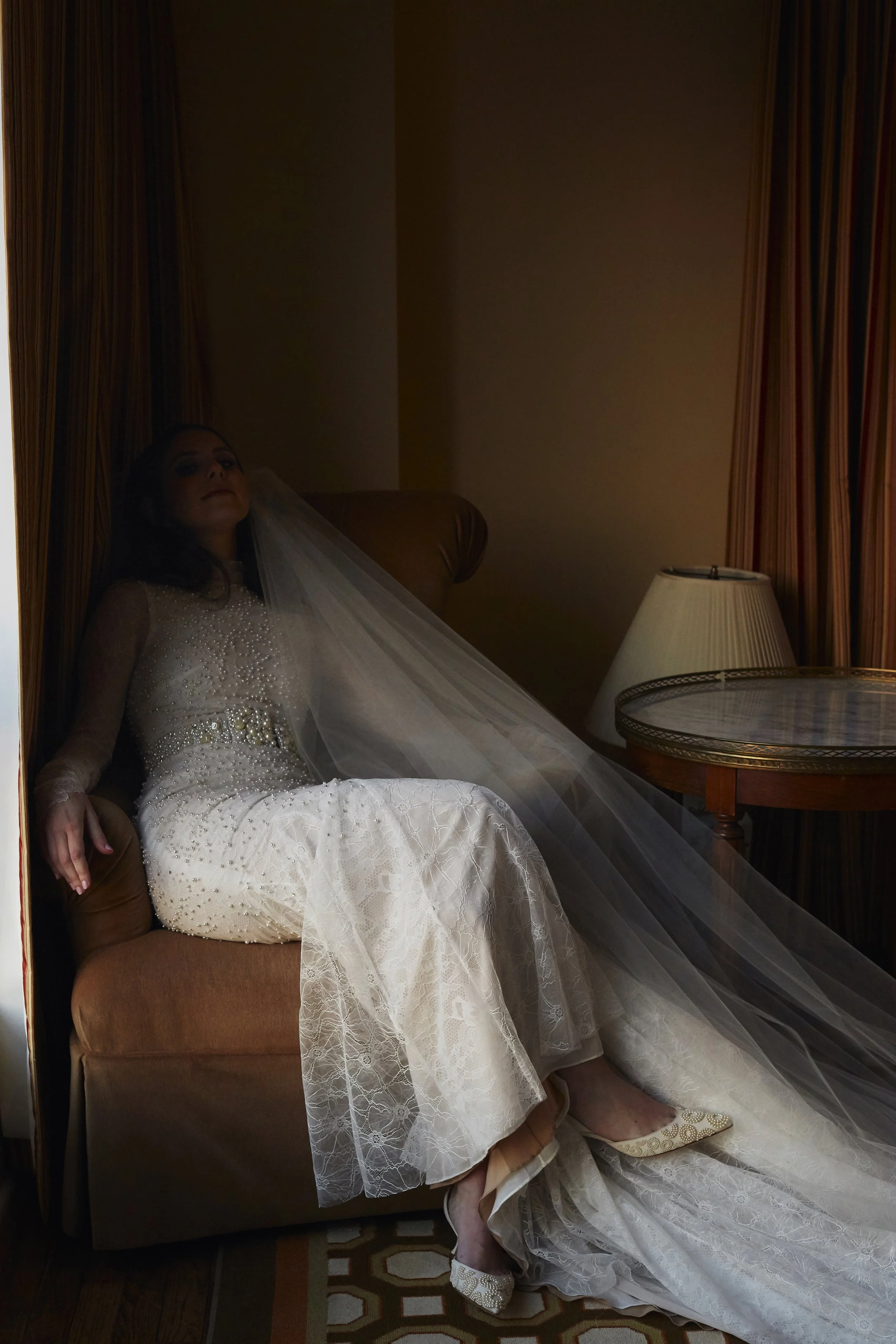 Woman in a wedding dress sitting on a tan armchair with a bridal veil, in a room with dark curtains and a table lamp.