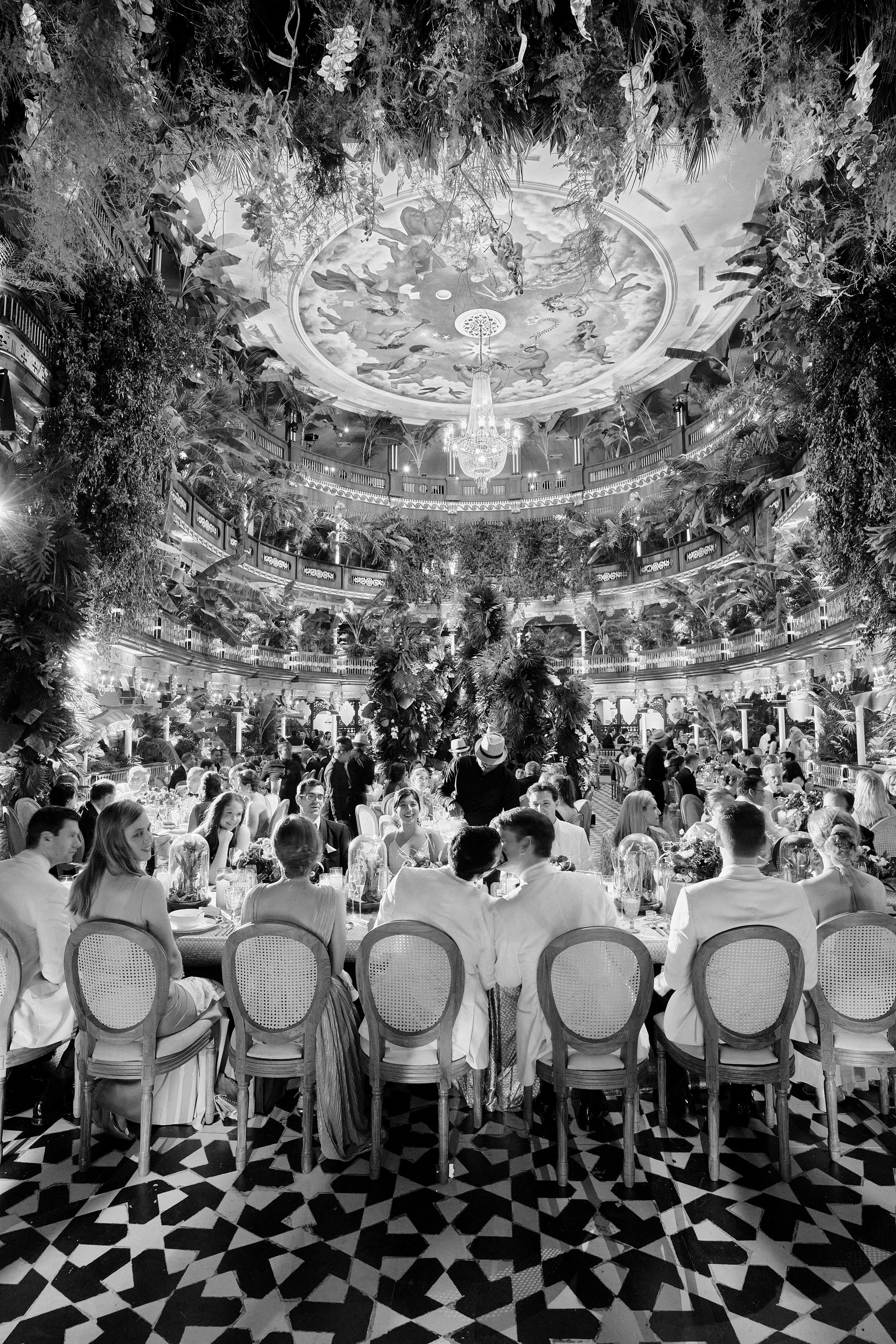 Elegant banquet hall filled with people seated at tables, lush greenery and plants adorning multiple levels, a painted ceiling with cherubs, and a large chandelier hanging from the decorated ceiling.