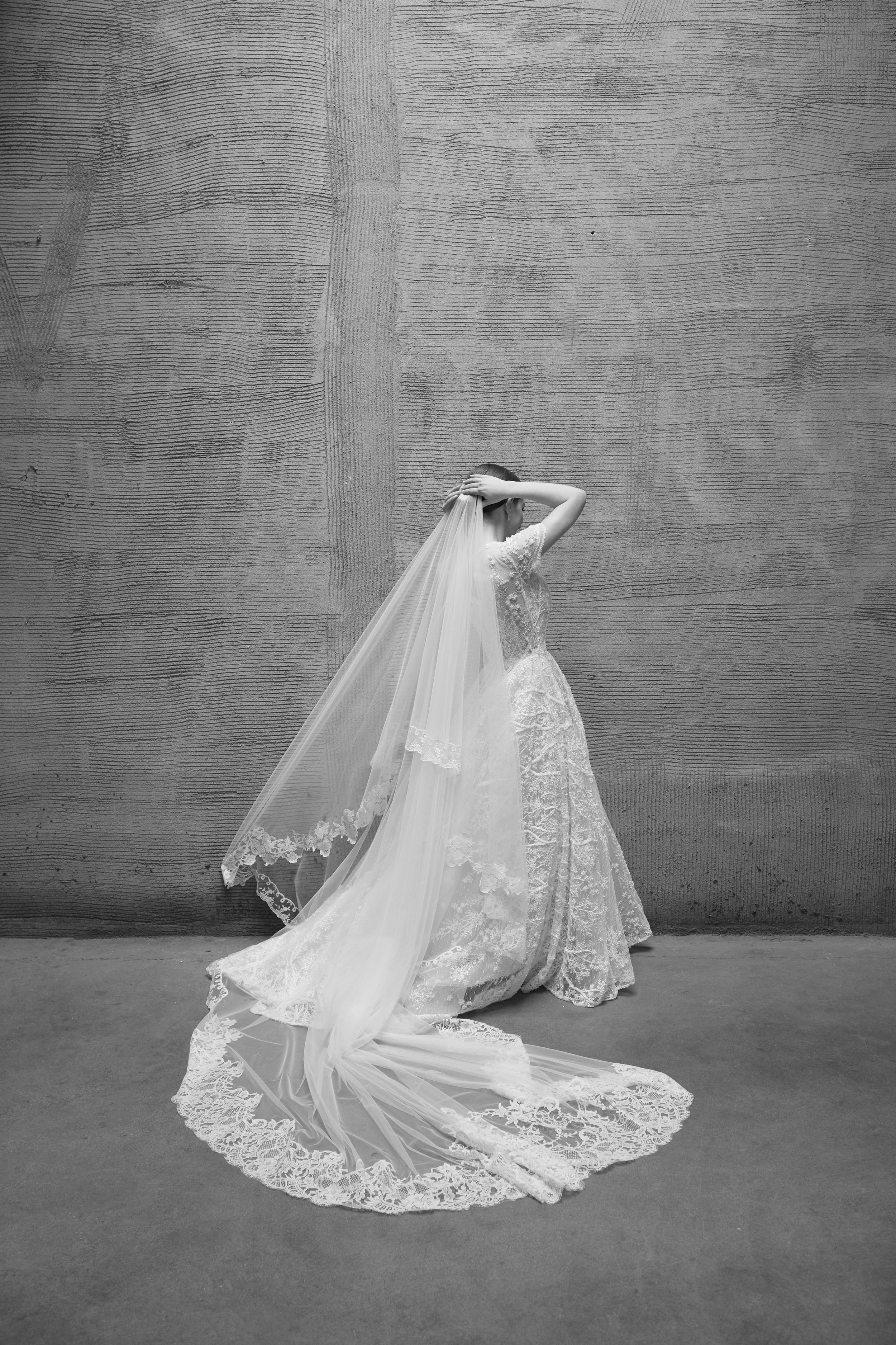 A woman in a wedding dress stands against a wooden wall, with her hand on her head and a lengthy, lace-trimmed veil flowing to the floor.