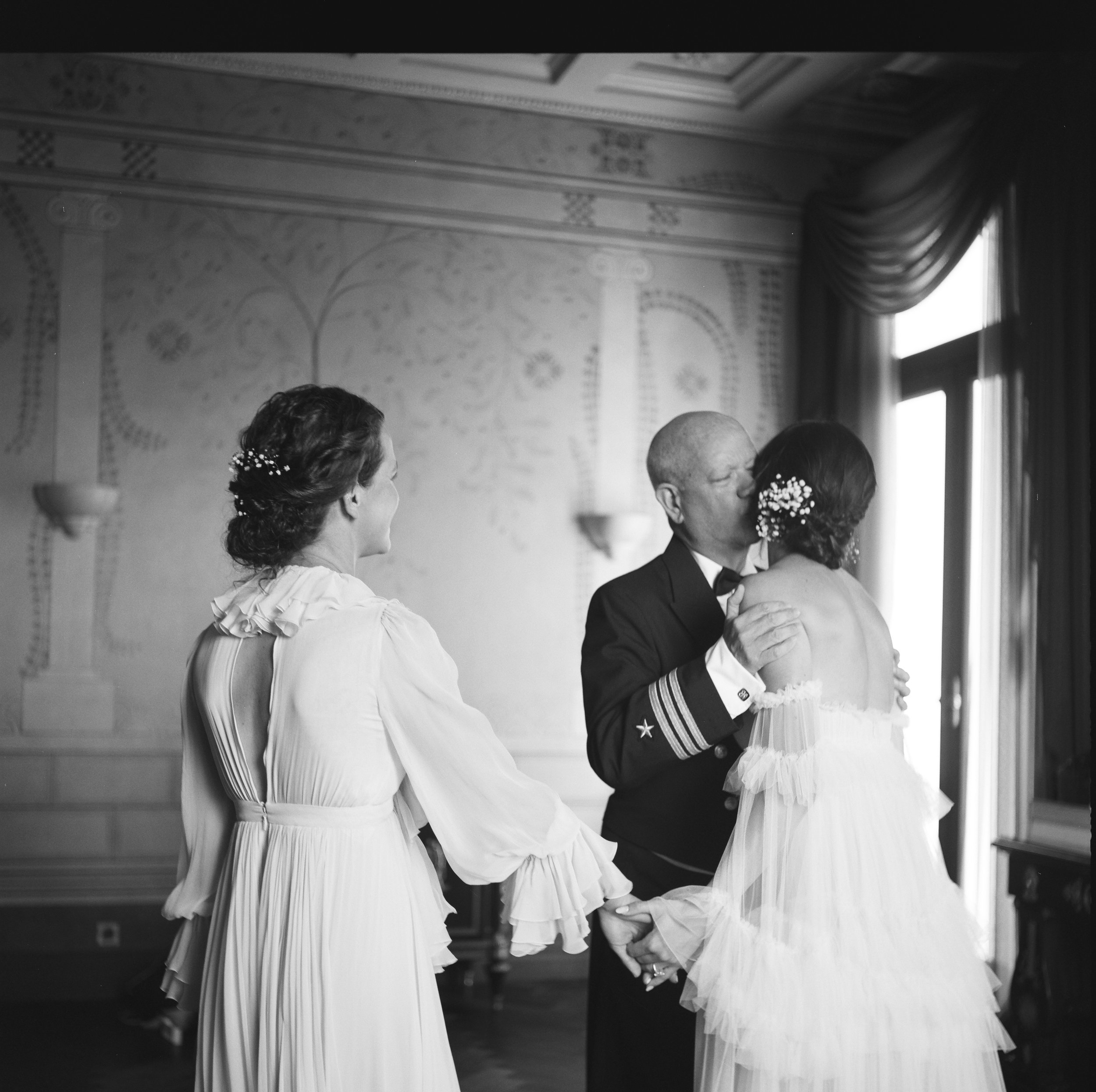 A black-and-white photograph of a wedding ceremony showing multiple couples dancing. The women are wearing elegant dresses with ruffled sleeves and flowers in their hair, while the men are dressed in suits. The room has ornate walls and a window with curtains.