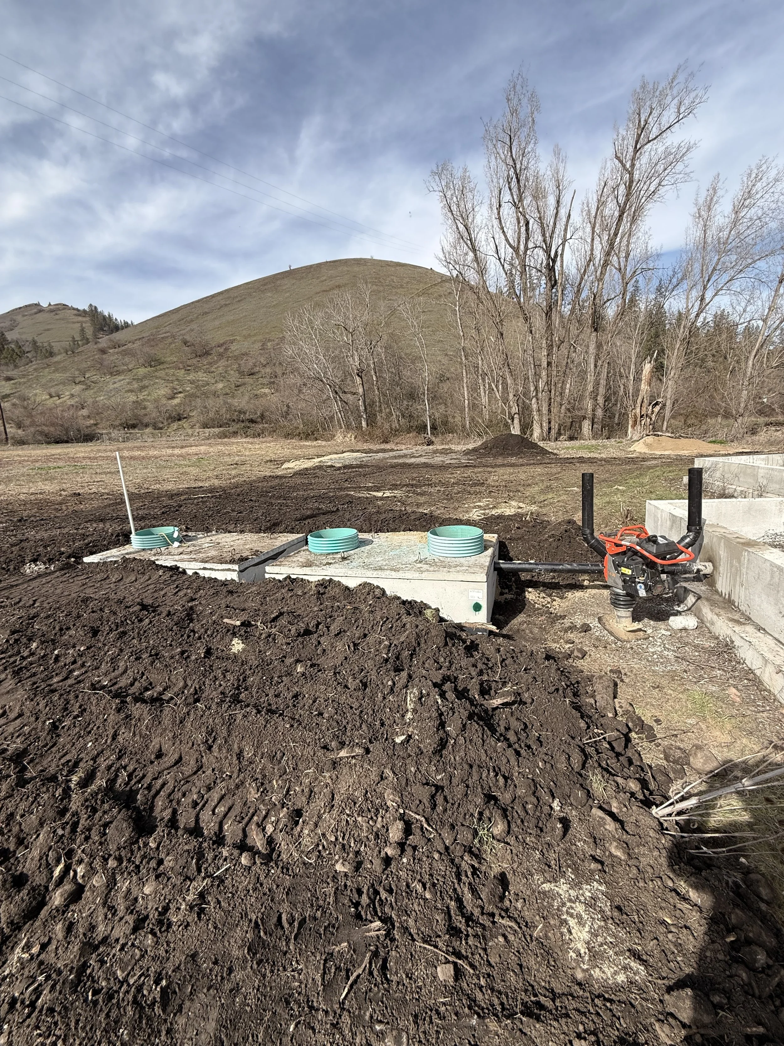 Construction site with freshly dug dark soil, concrete structures, and a power tool, set against a background of hills, trees, and a partly cloudy sky.
