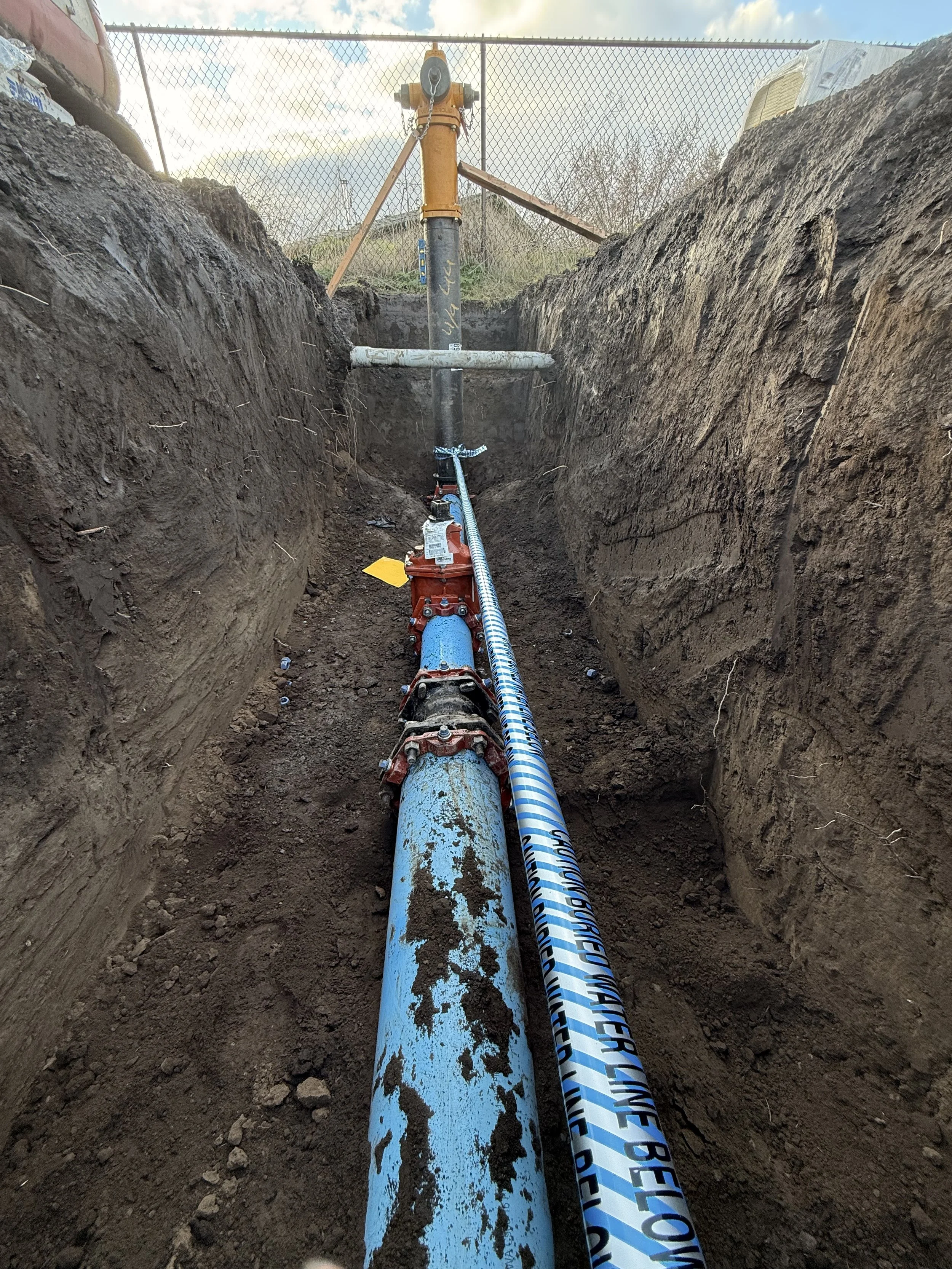 A construction site showing a trench with a blue pipe and an orange pipe in a dirt excavation. The pipes are connected with a metallic coupling, and there is a fire hydrant fitting on the blue pipe. The trench is flanked by vertical dirt walls, and a chain-link fence is visible in the background with some equipment on top.