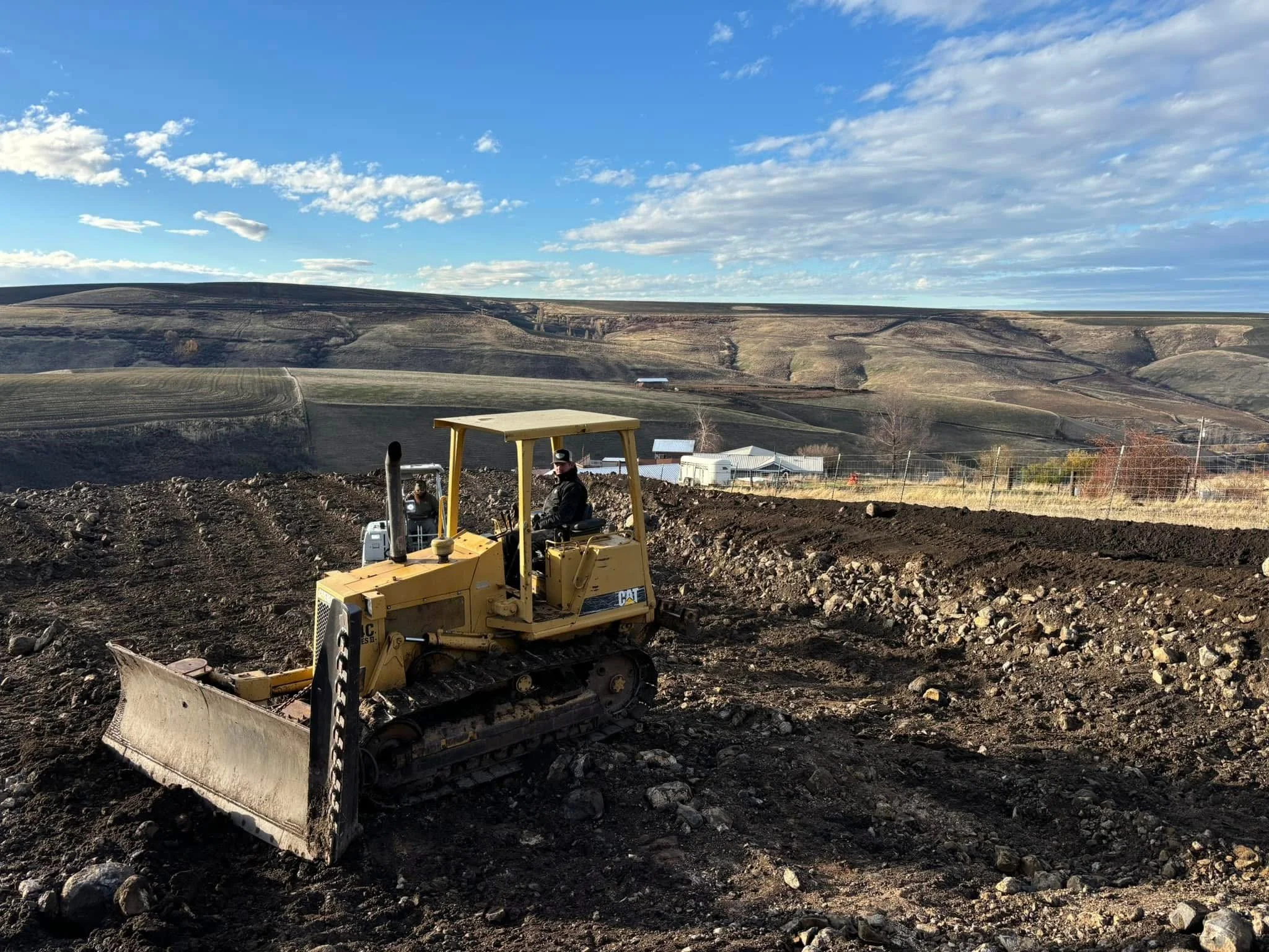 A person operating a yellow bulldozer on a construction site with a scenic landscape of rolling hills and blue sky with clouds in the background.