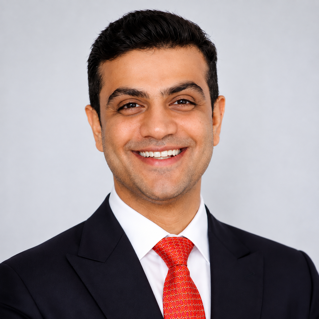 Headshot of a smiling man in a black suit, white shirt, and red tie against a light gray background.