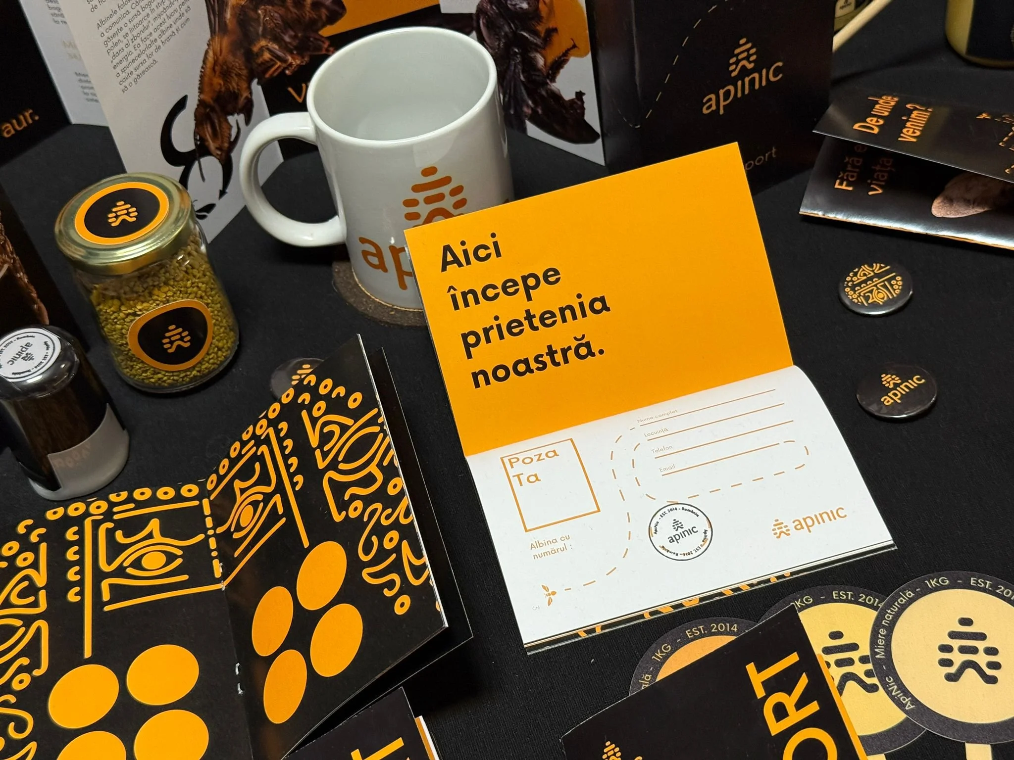 A display table with promotional items for Apinic, an herbal product brand. Items include a white mug with the Apinic logo, a jar of pellets, brochures, and informational cards with orange and black branding. There is a yellow sign with black text in Romanian that says 'Aici începe prietenia noastră'.